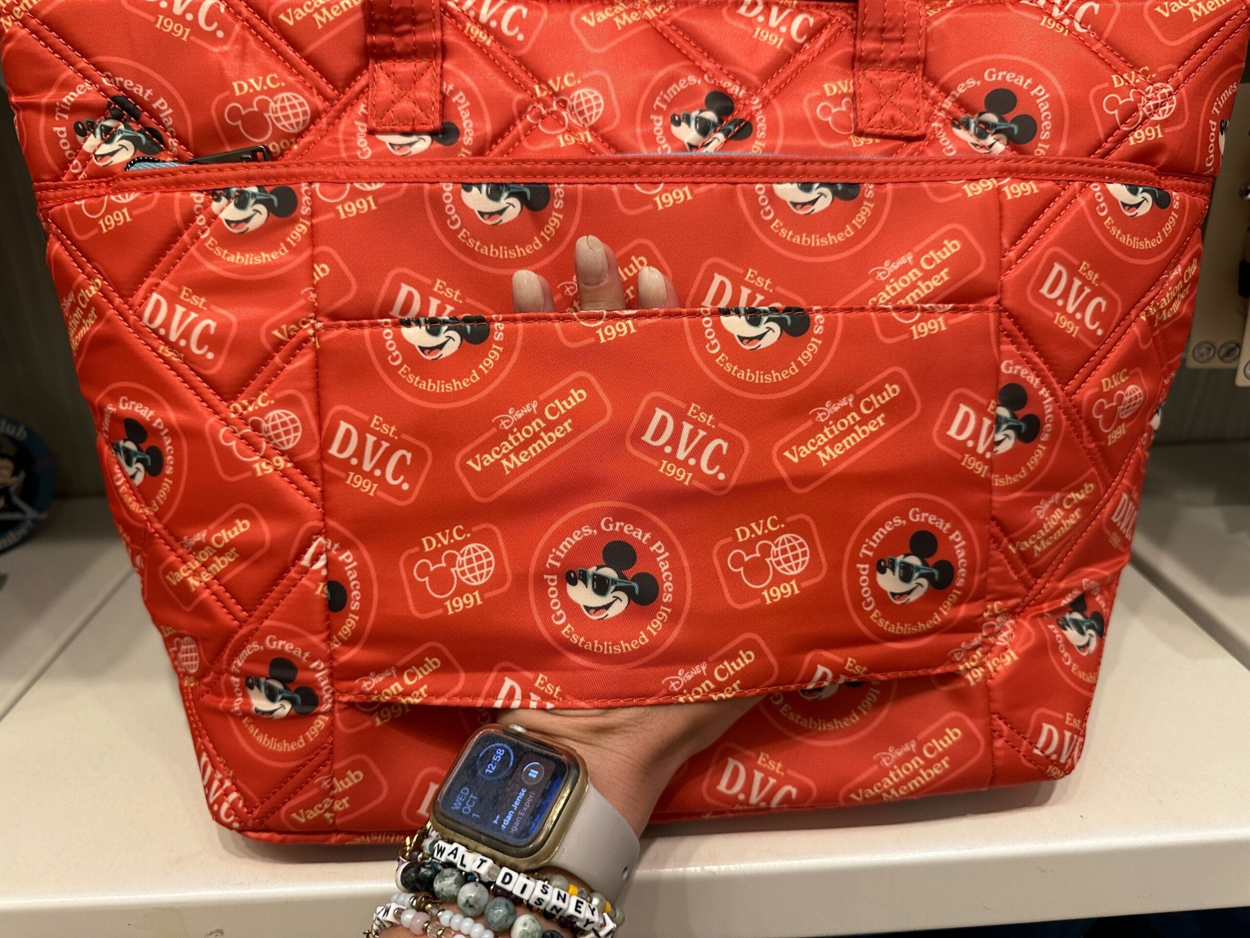 A hand holds a red Disney Vacation Club bag from the Merch Collection with Mickey Mouse patterns and an Apple Watch.