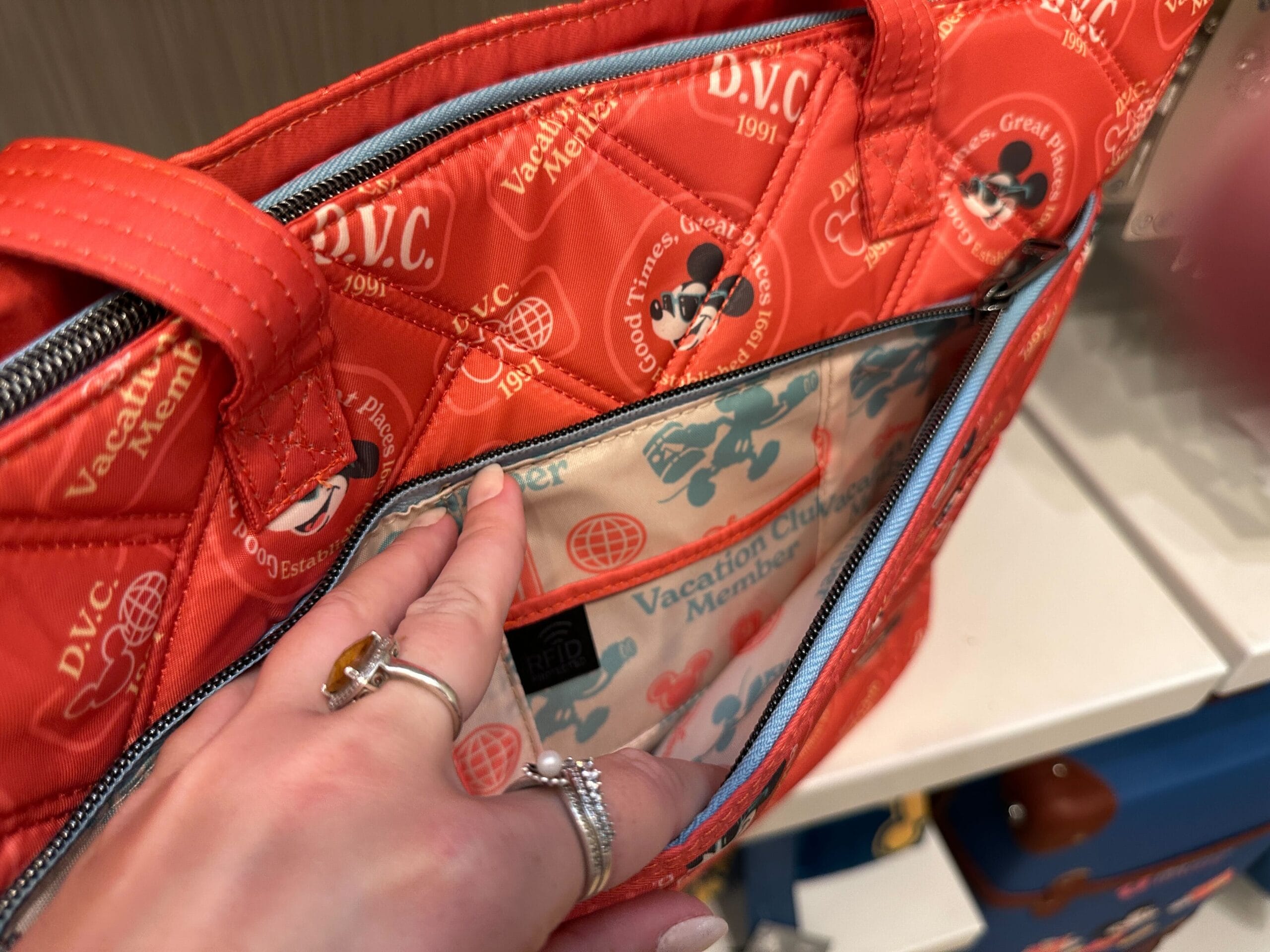 A hand opens a red Disney bag, revealing a Grand Floridian-inspired interior with DVC Member Merch and Mickey Mouse designs.