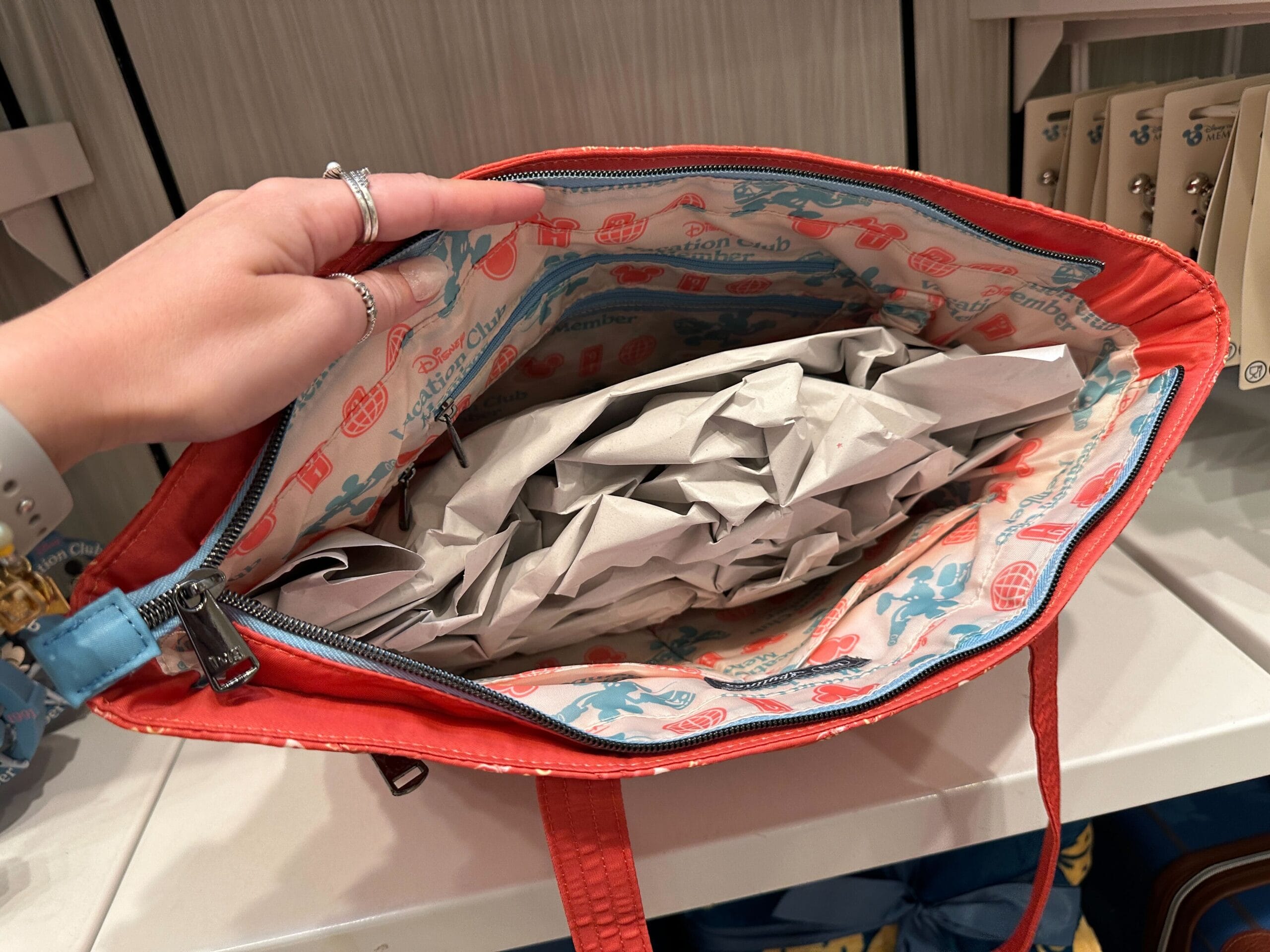A hand opens a red bag revealing crumpled paper and a printed lining from the Grand Floridian New Collection.