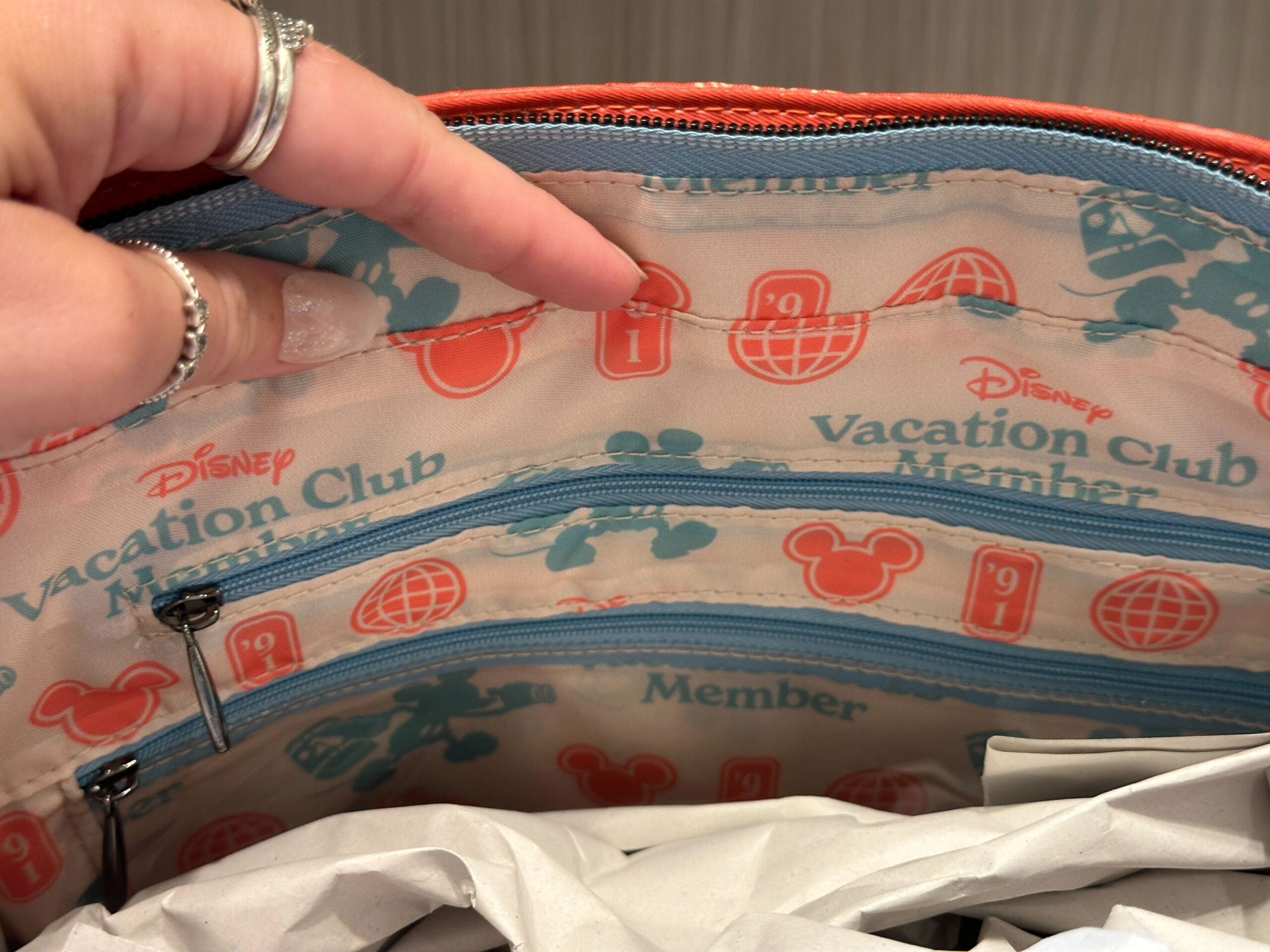 A hand holds open a Grand Floridian DVC Member-themed bag with blue zippers and red and blue printed icons inside.