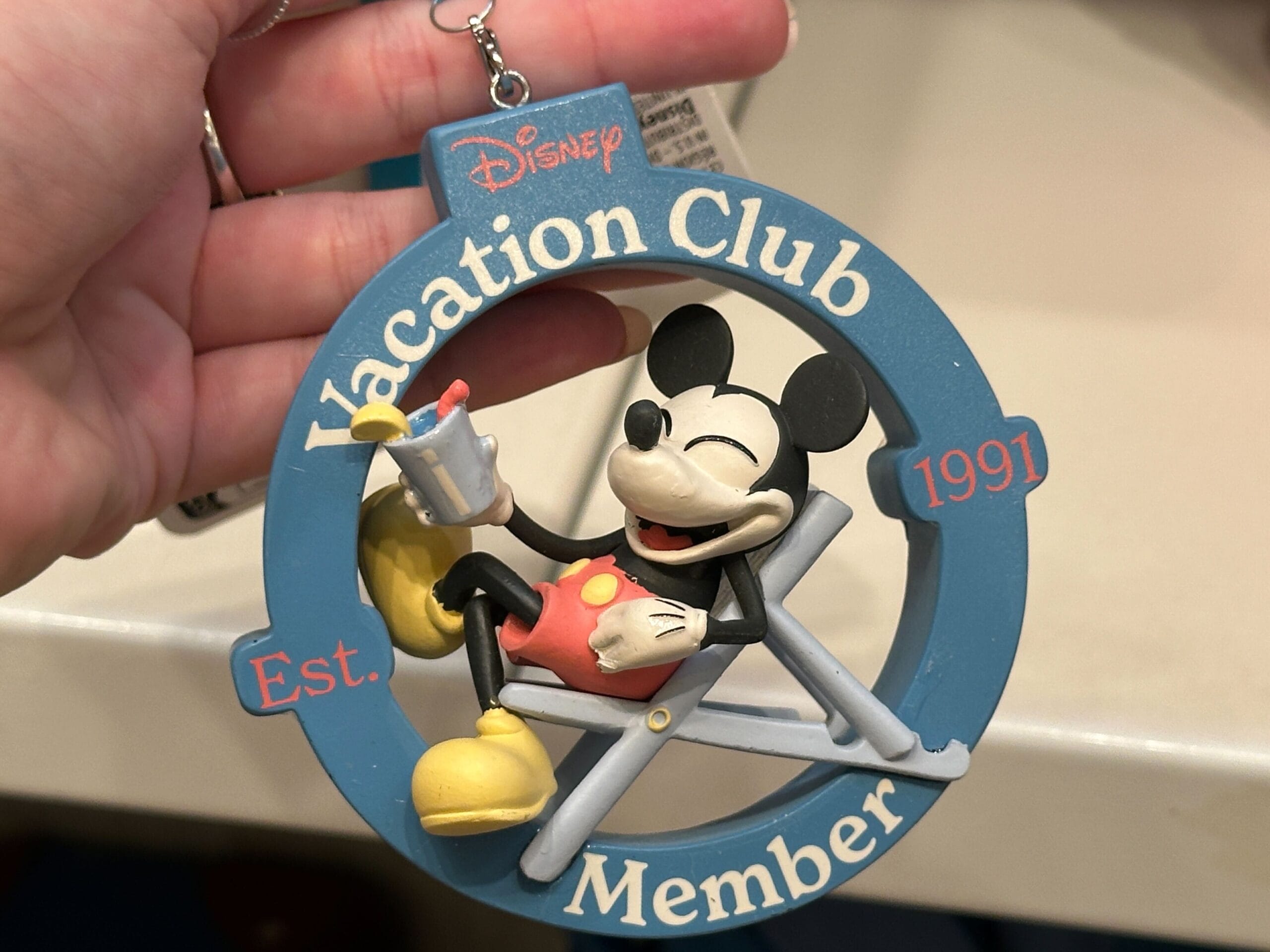 A hand holds a DVC Member ornament of Mickey Mouse lounging, part of the Grand Floridian Merch Collection.