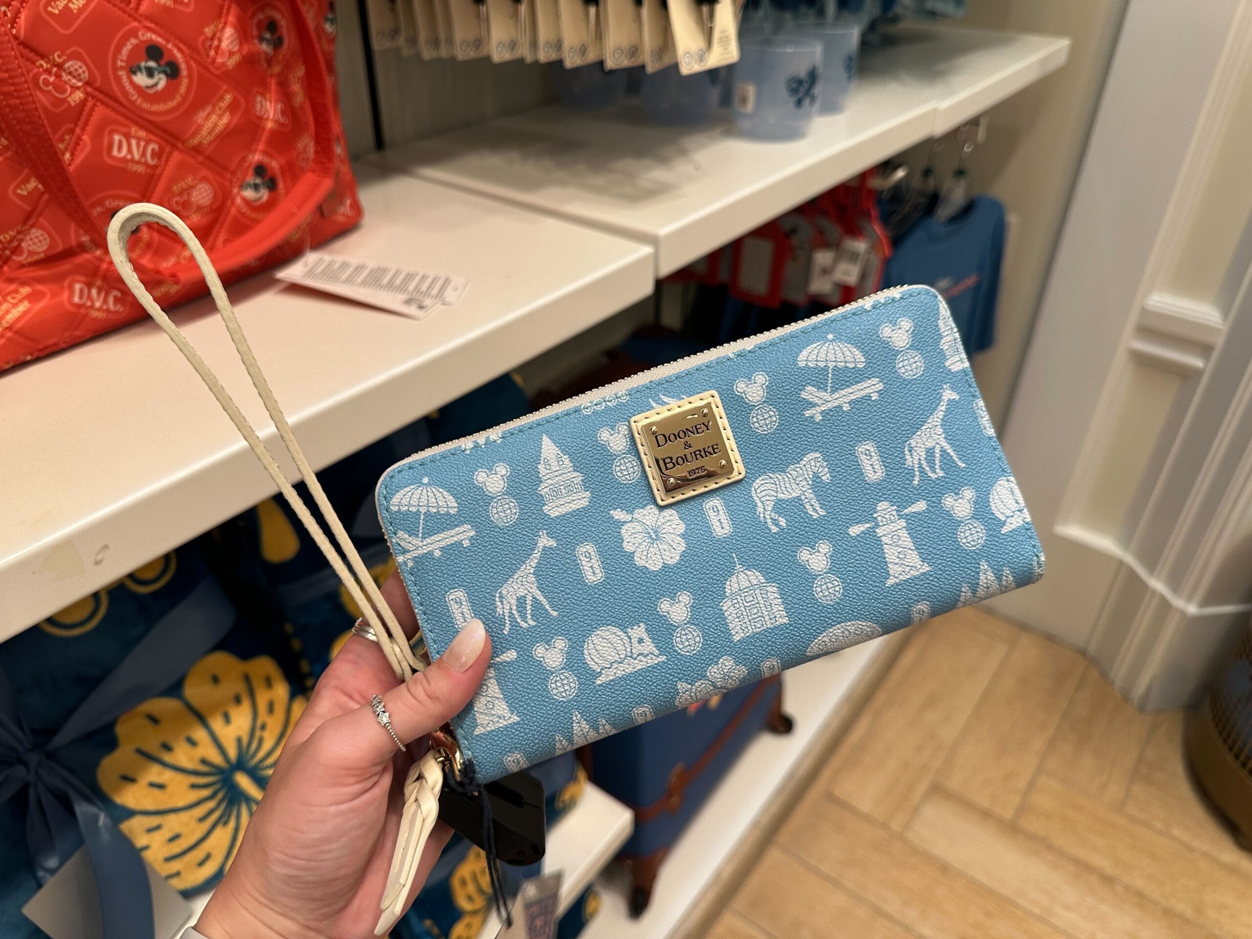 A hand holds a blue Disney Dooney & Bourke wallet from the new collection with white safari patterns in a store.