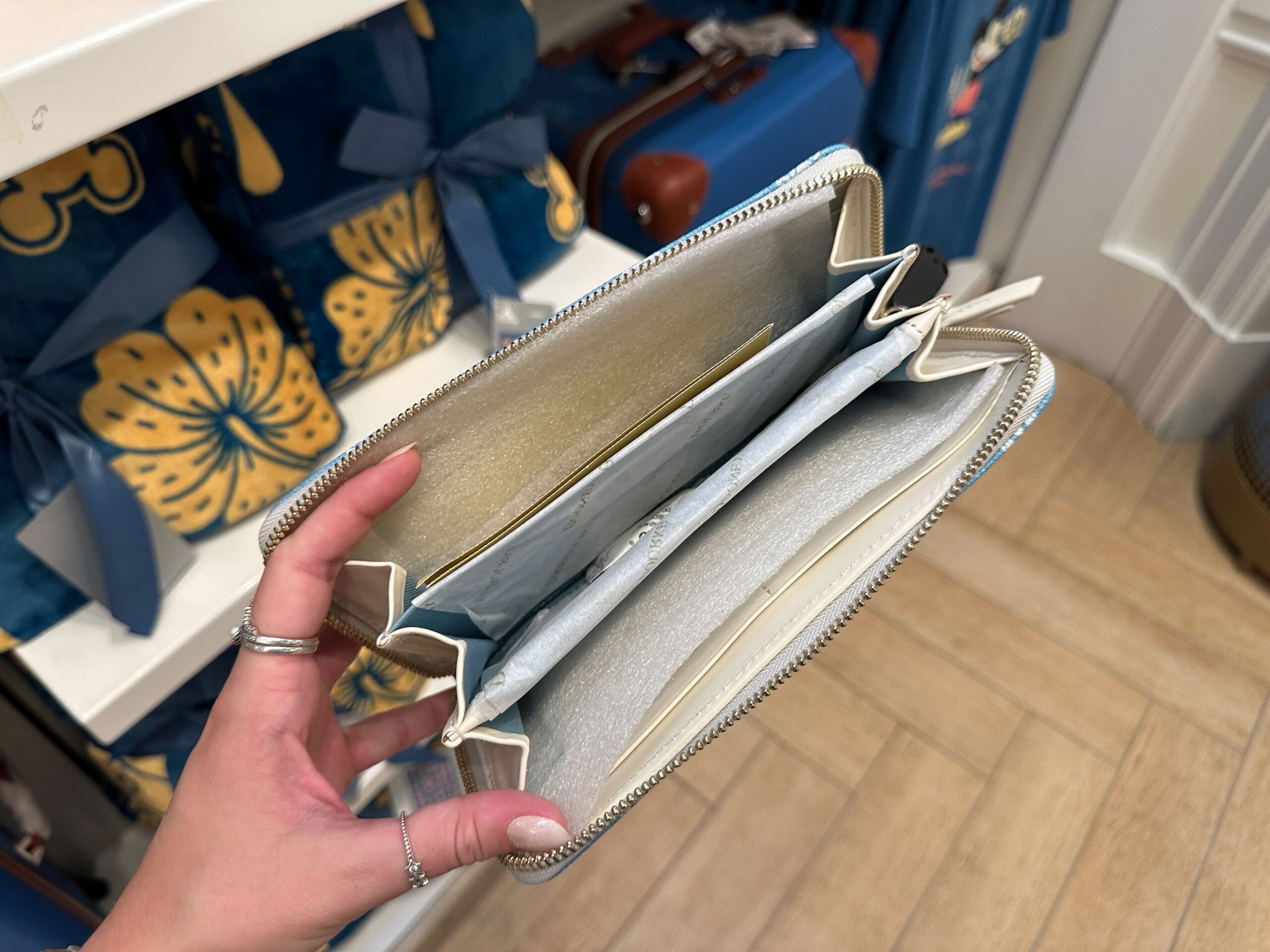A hand holds open an empty zippered wallet in a store with Grand Floridian merch collection bags on the shelf.
