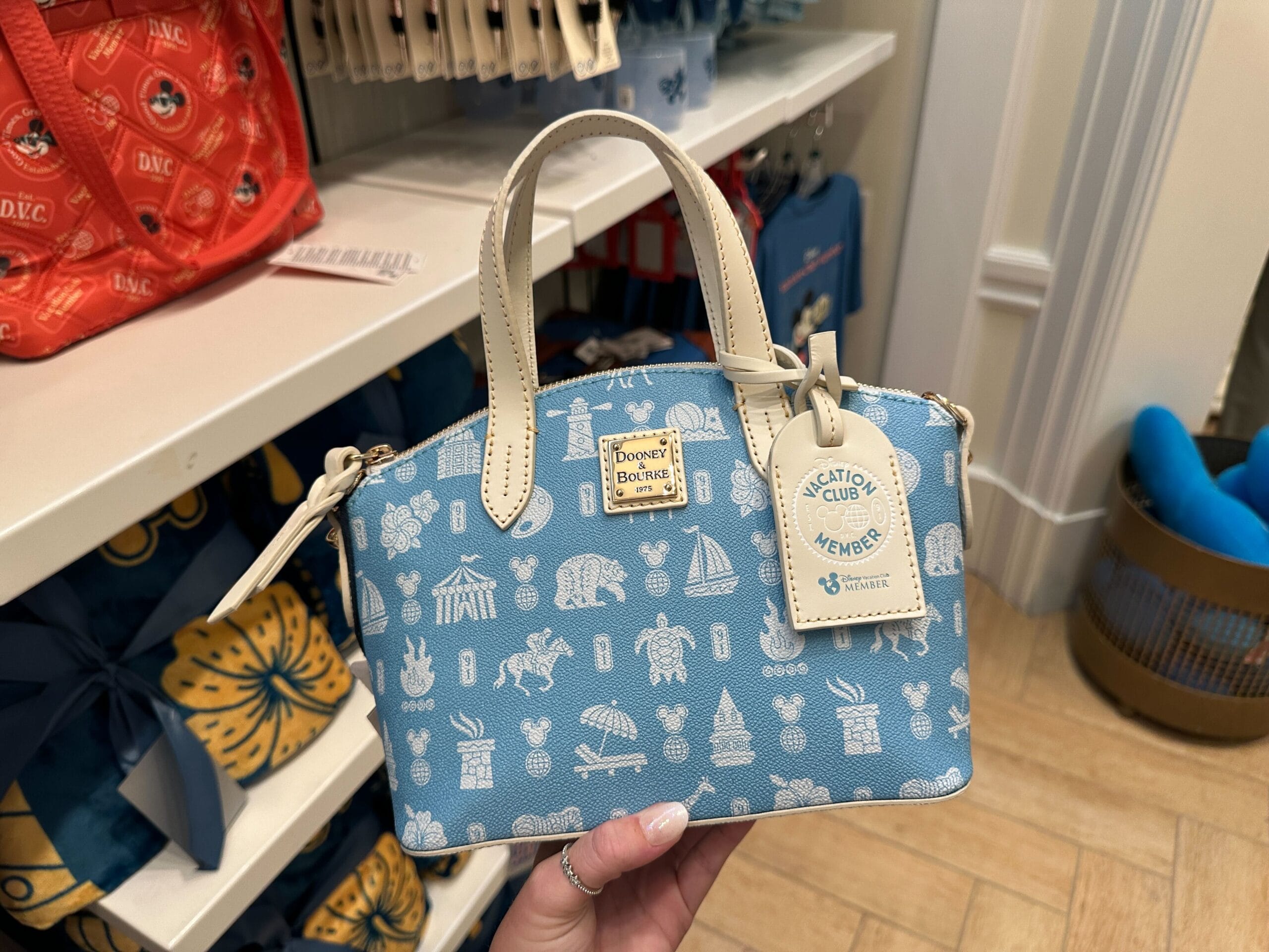 A hand holds a blue Dooney & Bourke DVC Member Merch handbag with Disney icons in a Grand Floridian store display.