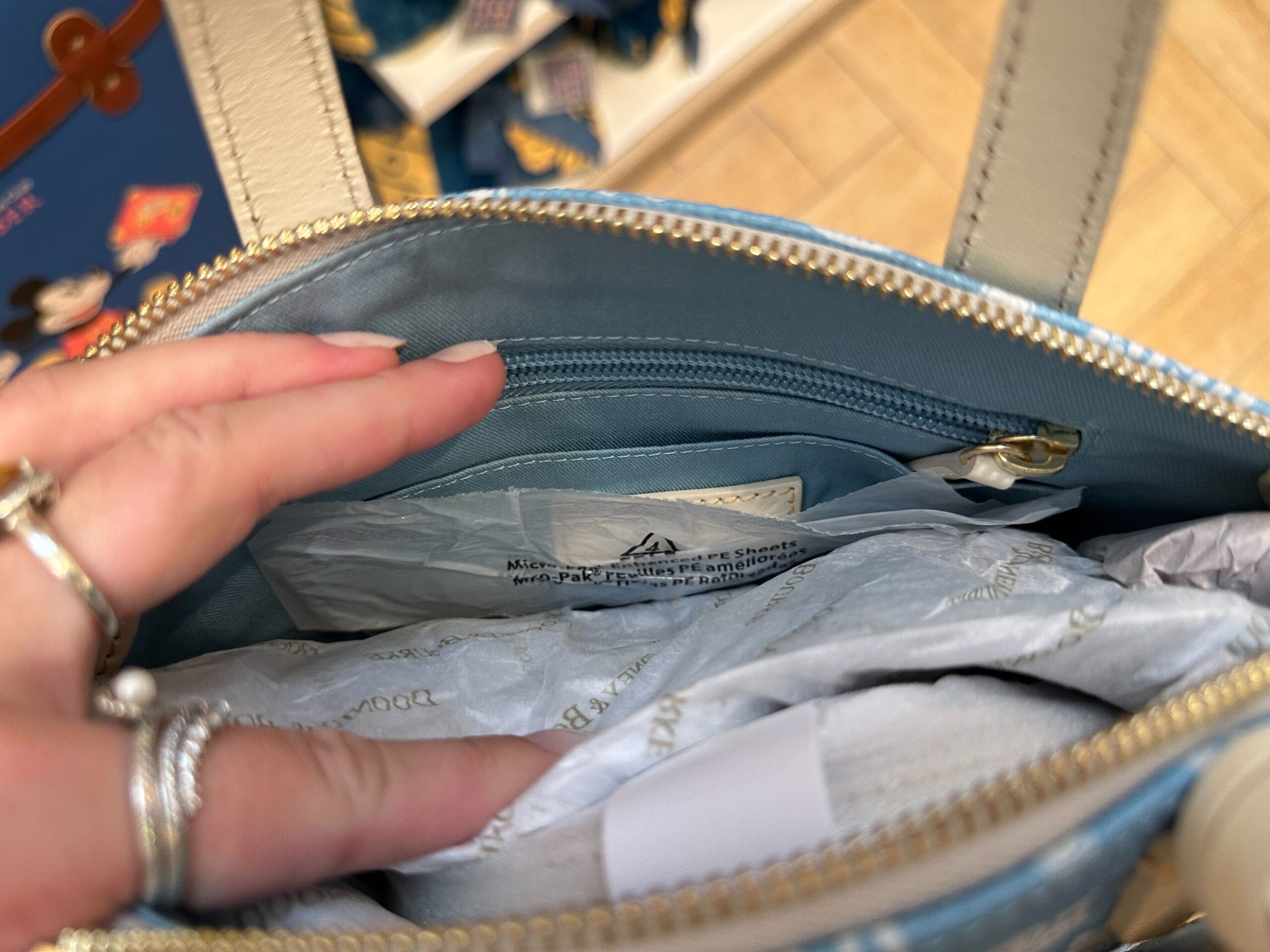 A hand opens a light blue purse from the Grand Floridian Merch Collection, showing tissue paper and a zippered pocket inside.