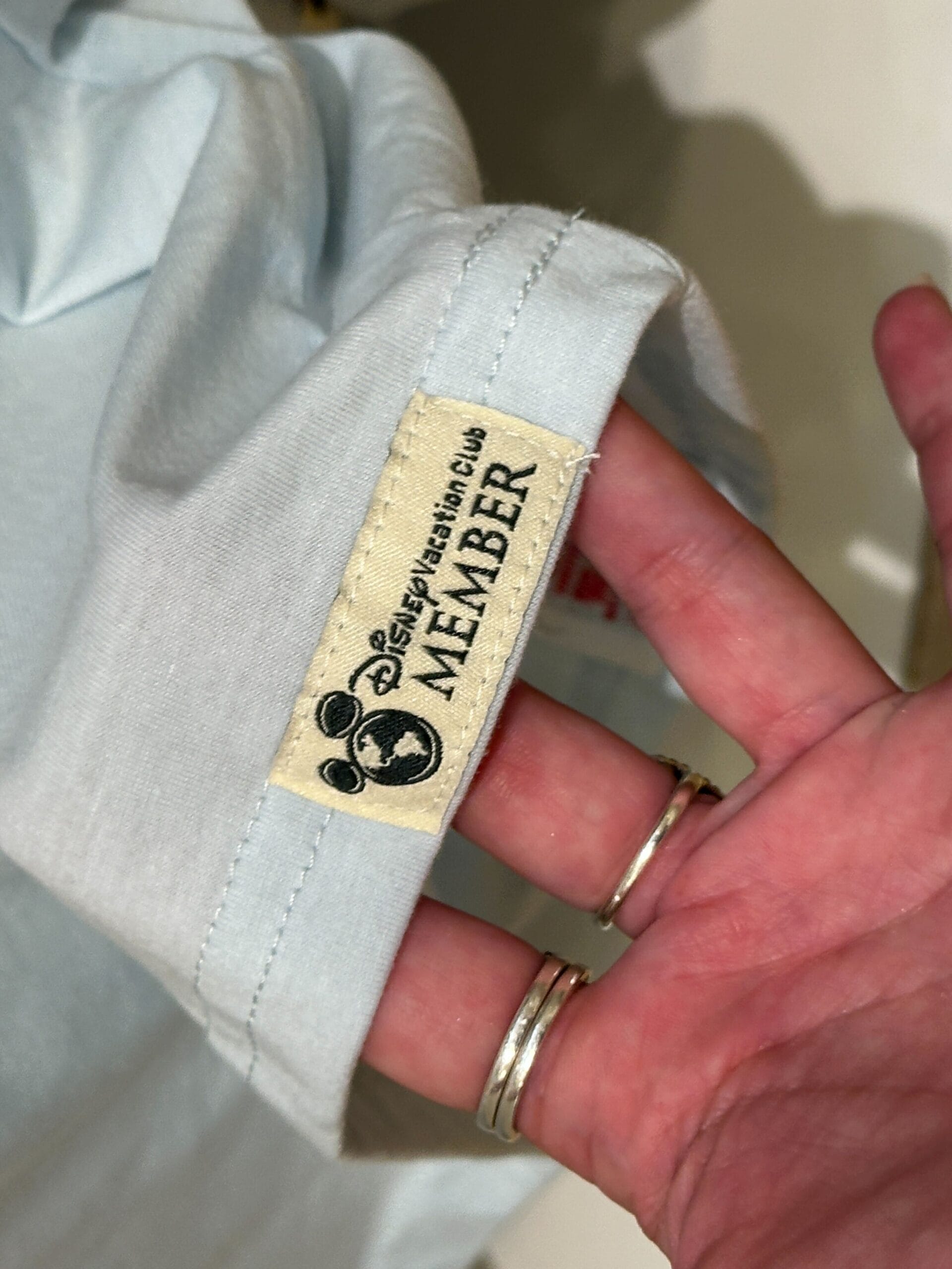 A hand holds Grand Floridian DVC Member Merch: a light blue shirt with a "Disney Vacation Club MEMBER" Mickey tag.