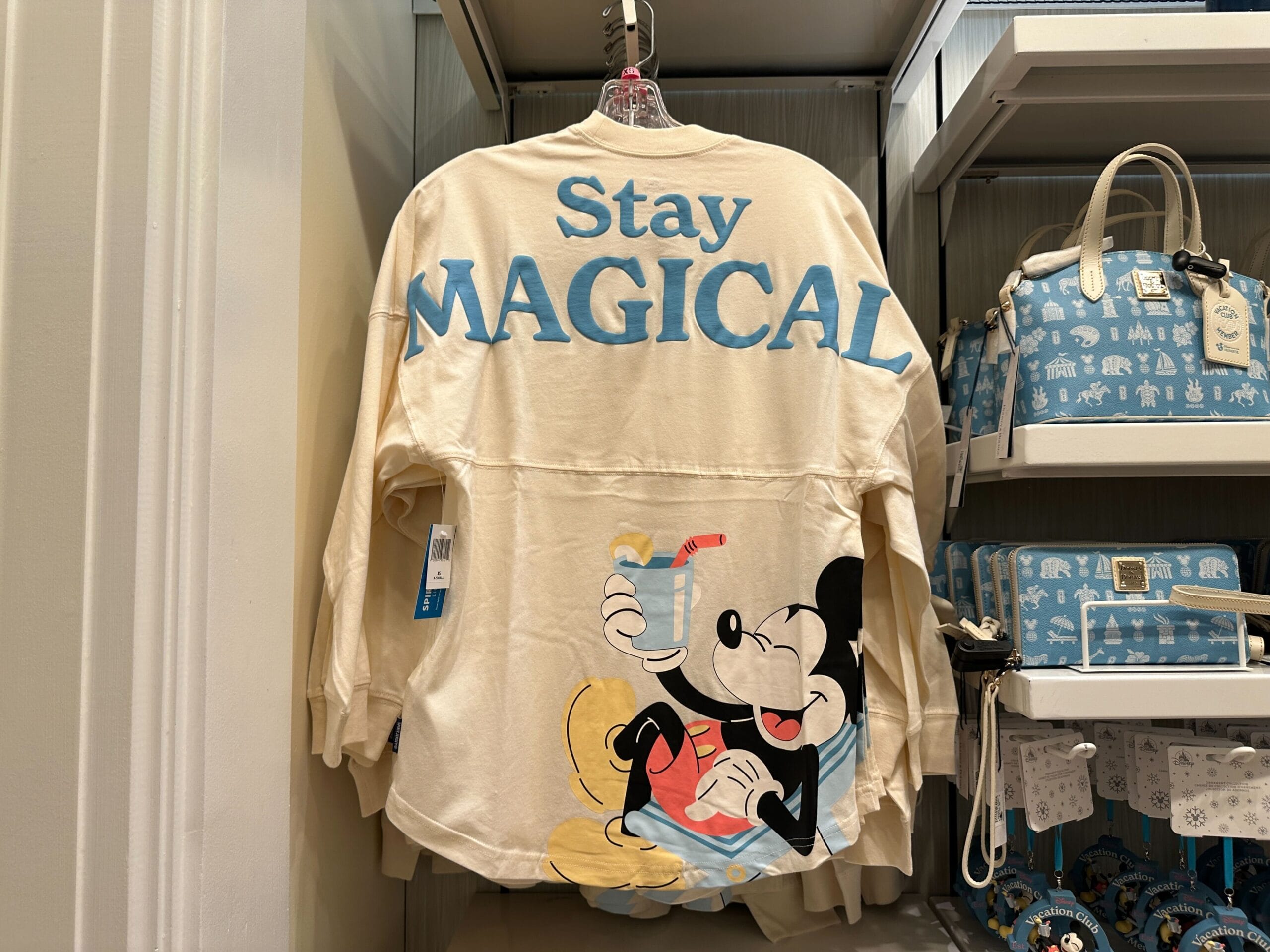 Back of Disney Vacation Club Member-exclusive cream Spirit Jersey shirt with blue "Stay Magical" text and Mickey Mouse relaxing on a beach chair.