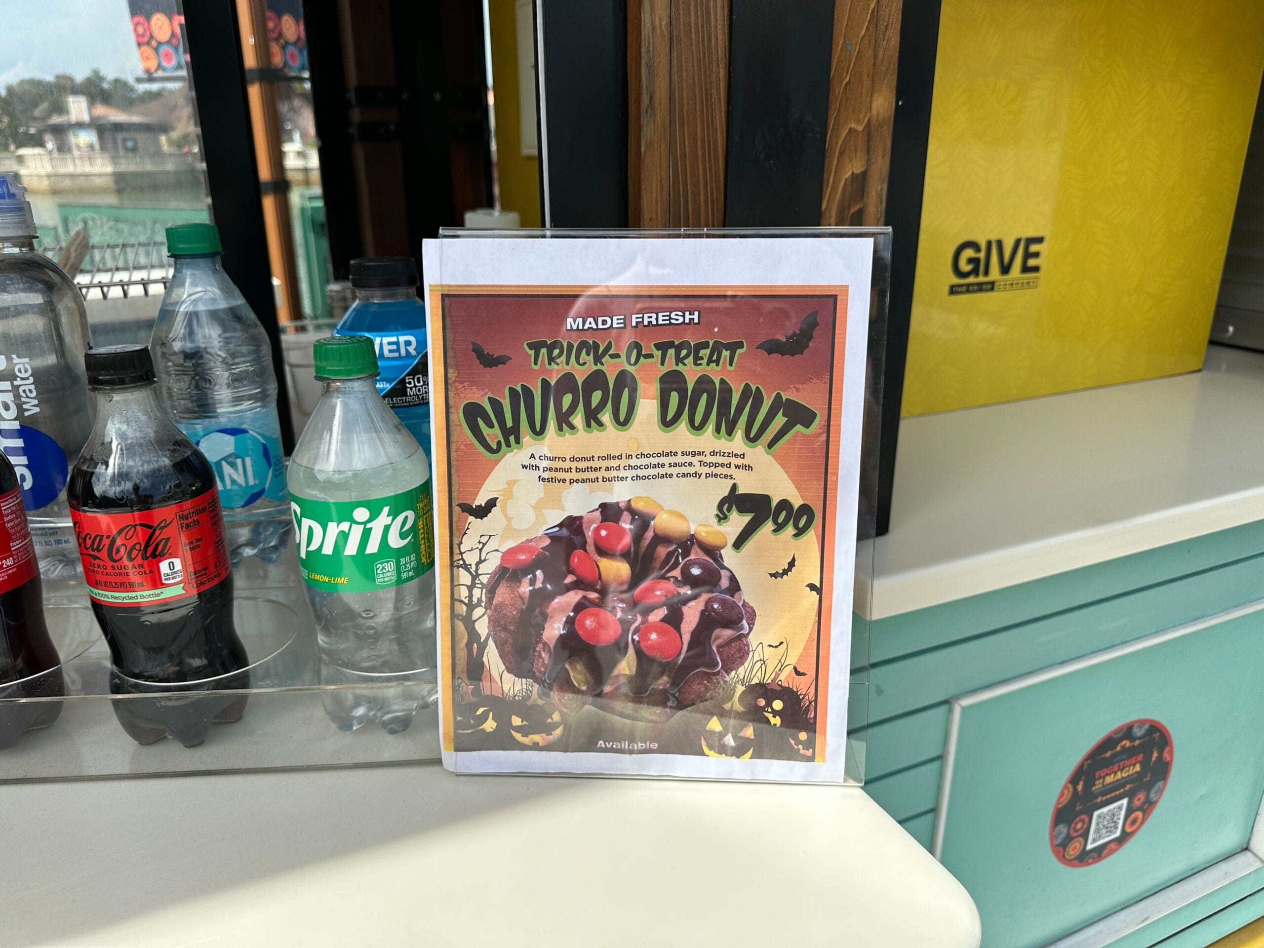 Sign advertising a "Trick-O-Treat Churro Donut" for $7.99, surrounded by soda bottles and a yellow box.