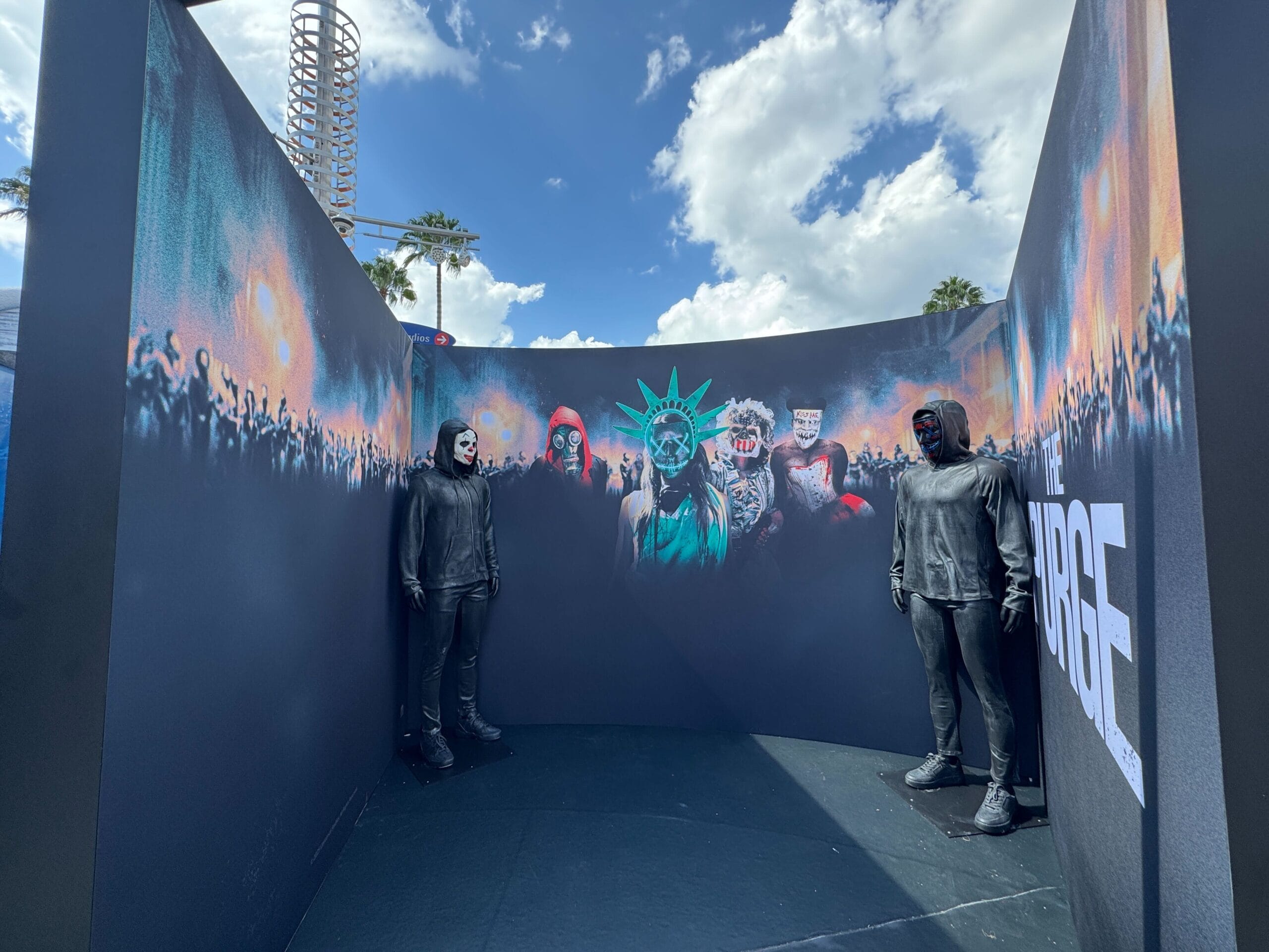 Three people in black costumes stand in a dark booth with "The Purge" mural at Universal CityWalk, blue sky above.