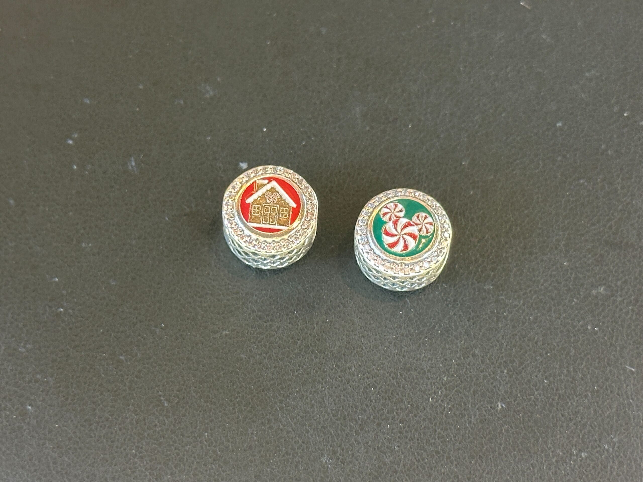 Two round sterling silver Pandora Charms on a black surface: one with a gingerbread house on a red background, the other with a Mickey head made of peppermint candies on a green background.