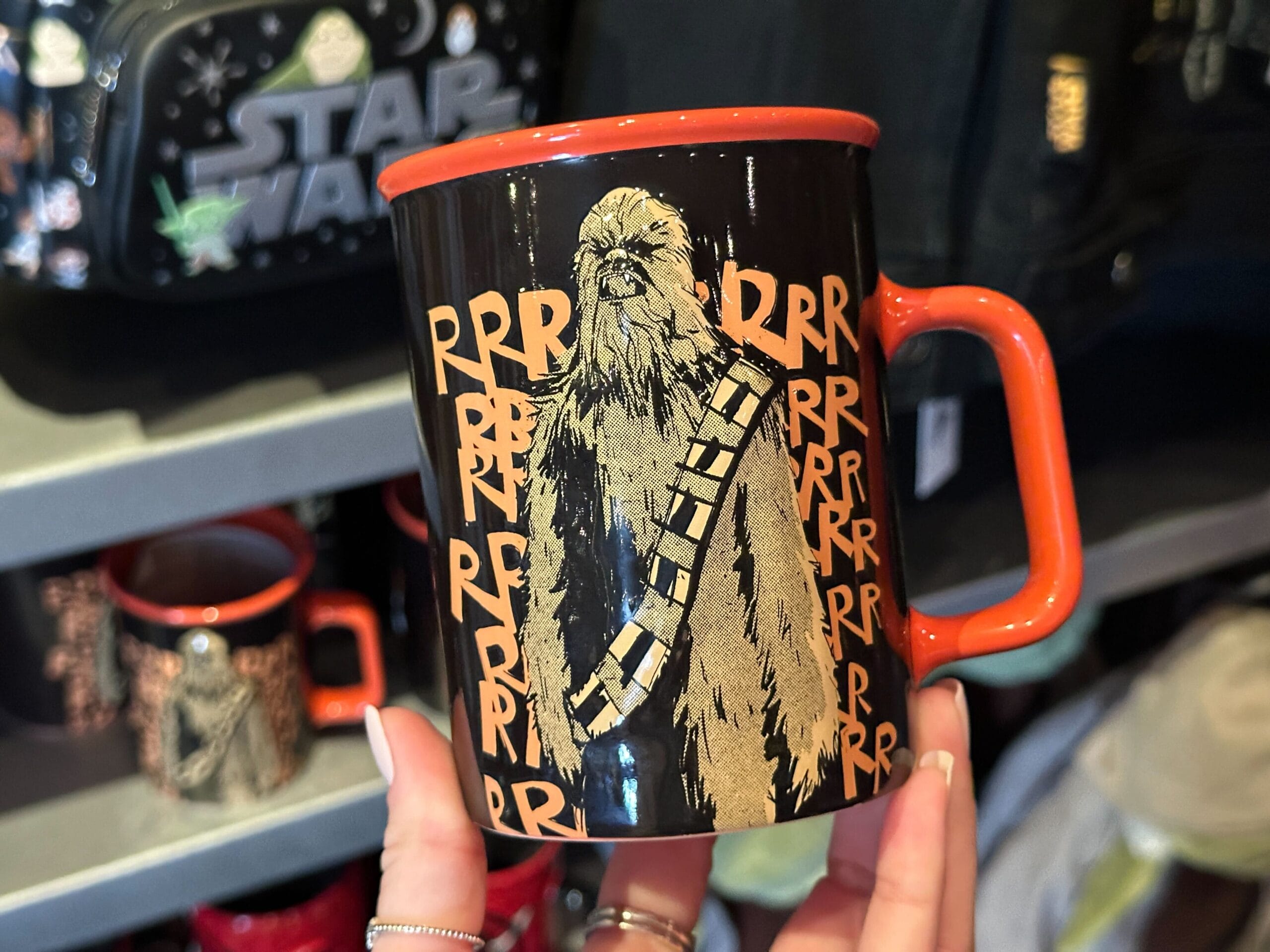 A hand holds a black Star Wars mug with Chewbacca in front of repeating "RRR" text and an orange rim and handle.