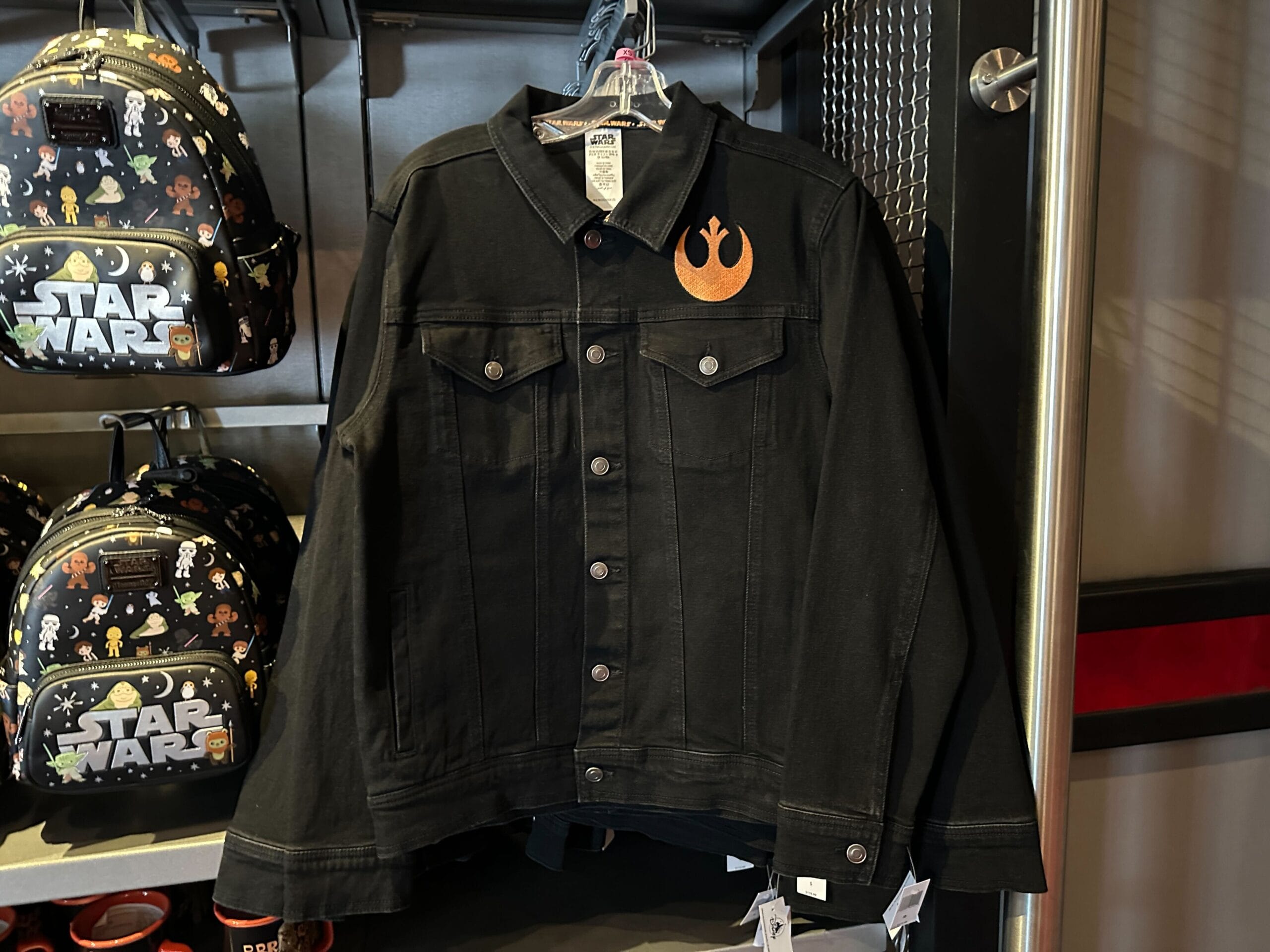 Black jean jacket with an orange Rebel Alliance Star Wars logo on the chest, hanging next to Star Wars-themed backpacks.
