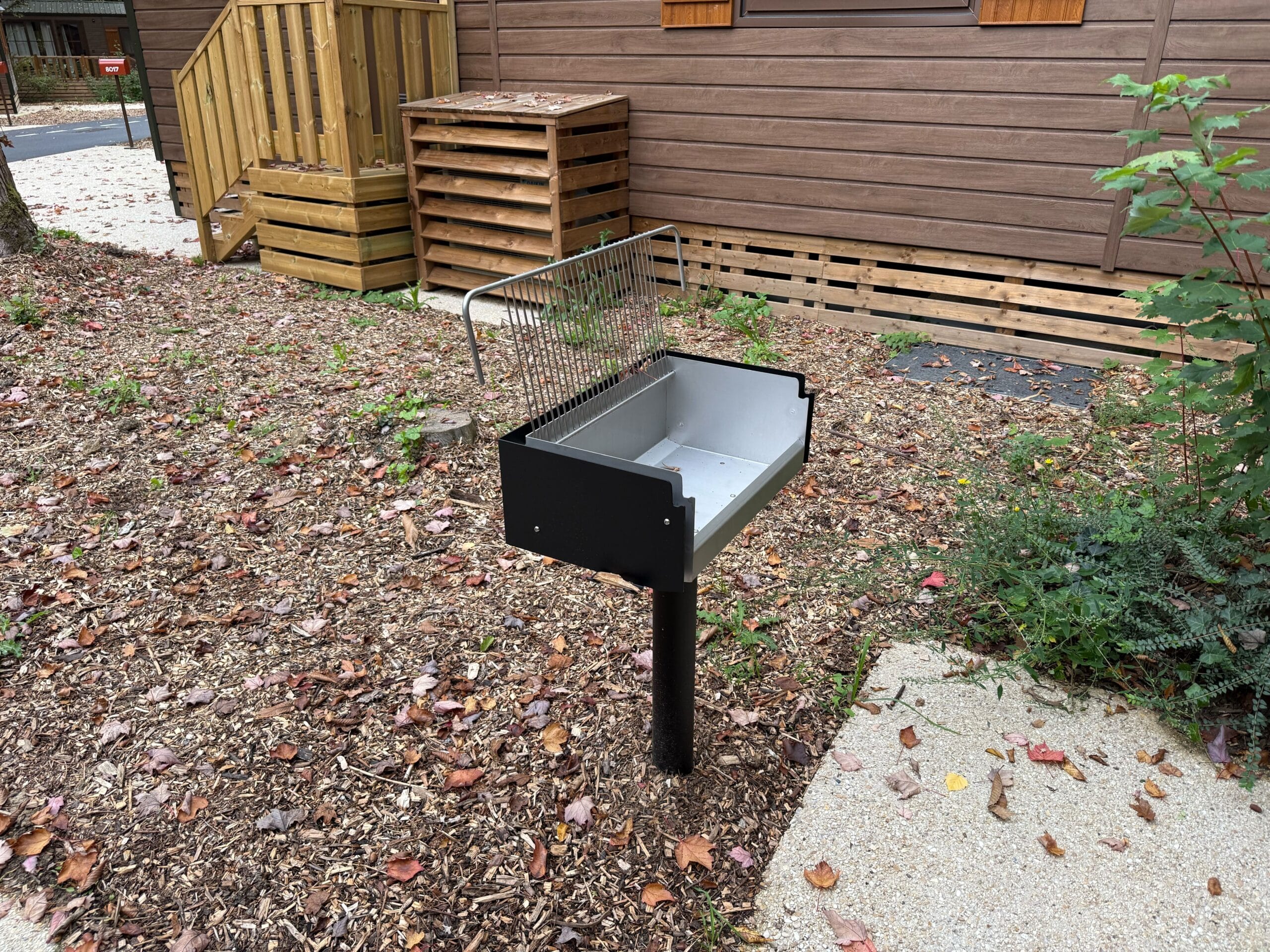 A metal outdoor grill on a pole stands outside a Davy Crockett Ranch cabin at Disneyland Paris, surrounded by mulch and leaves.