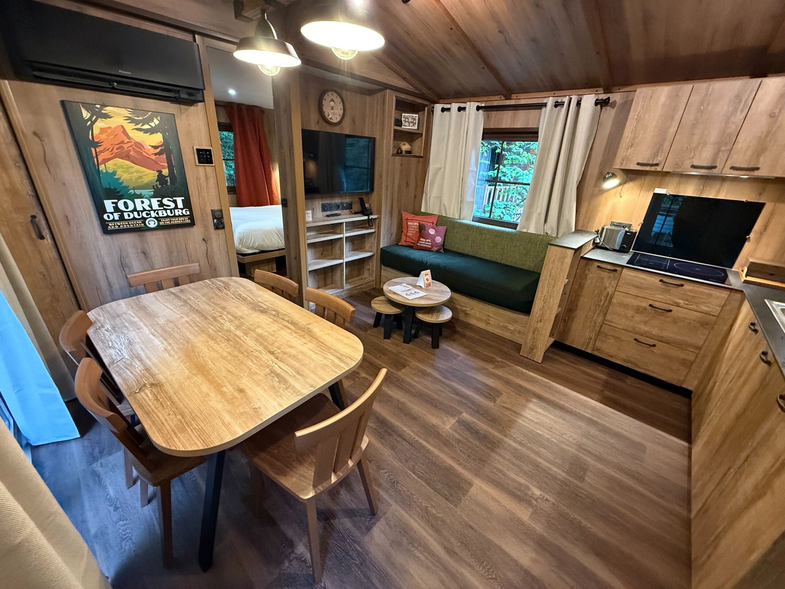 Charming Davy Crockett Ranch cabin room near Disneyland Paris, featuring a dining table, kitchenette, sofa, TV, and Forest of Duckburg poster.