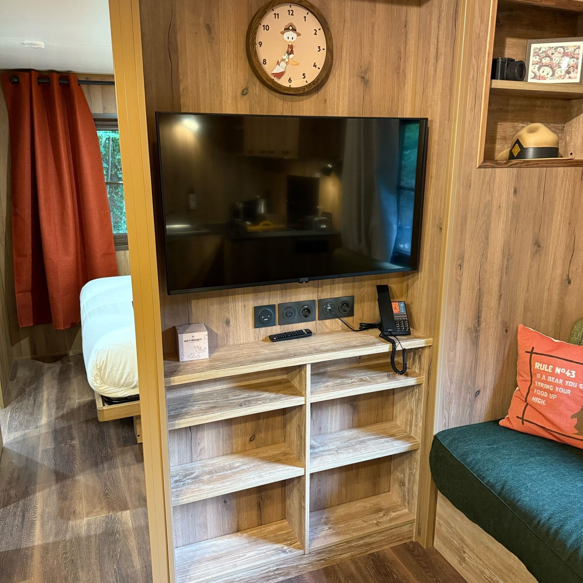 A wall-mounted TV sits beneath a clock in a cozy wood-paneled Cabin Room at Disneyland Paris’s Davy Crockett Ranch.