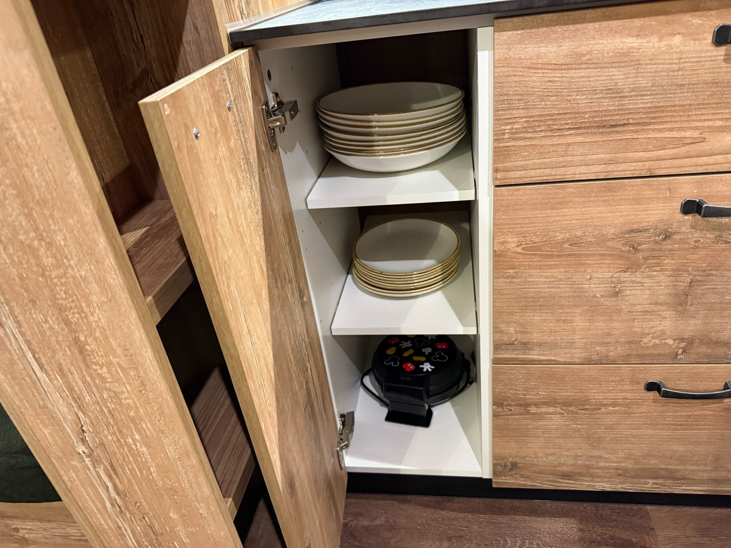 Open kitchen cabinet in a rustic Davy Crockett Ranch cabin at Disneyland Paris, displaying plates and a waffle maker.