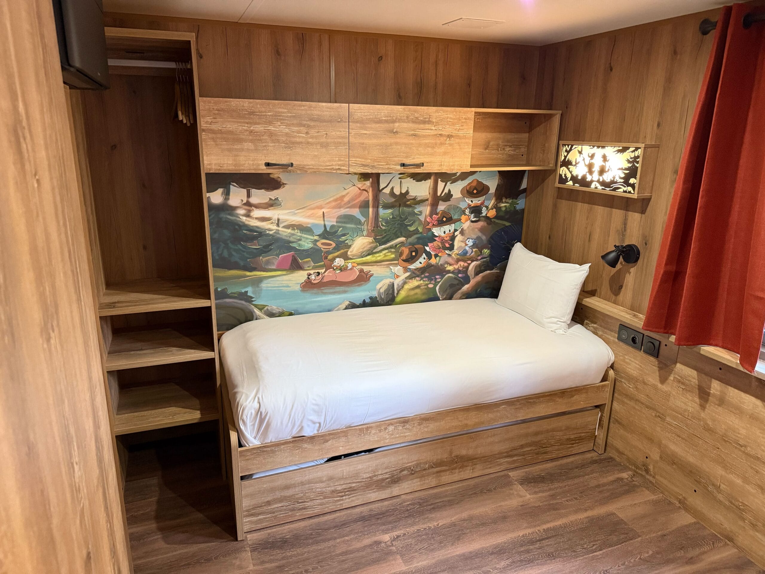 A cozy single bed with a white pillow, Junior Woodchucks mural, and red curtains in a Davy Crockett Ranch cabin at Disneyland Paris.