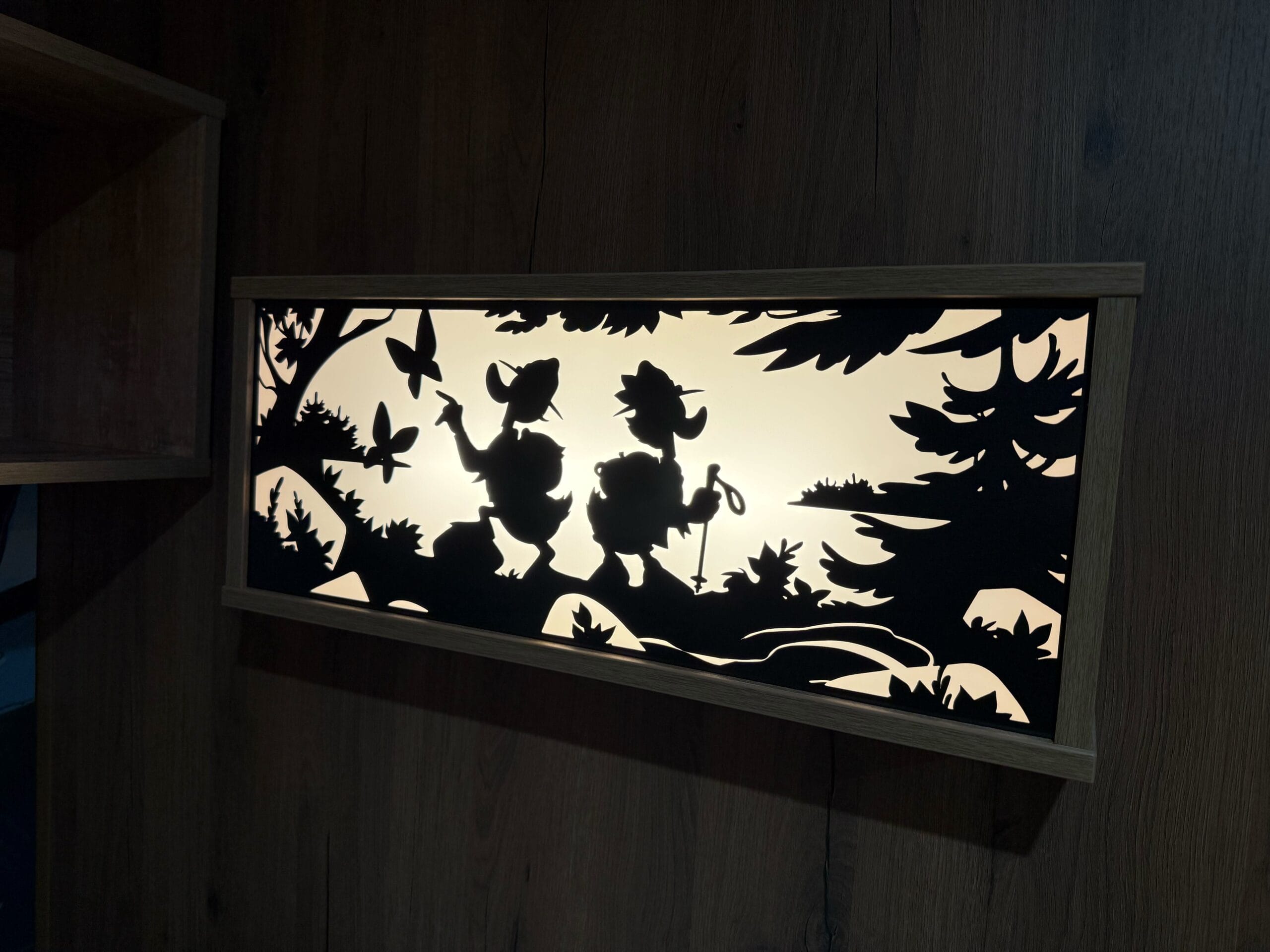 A rectangular light fixture with a cutout silhouetted scene of Donald and Daisy Duck on a hike surrounded by trees and butterflies.