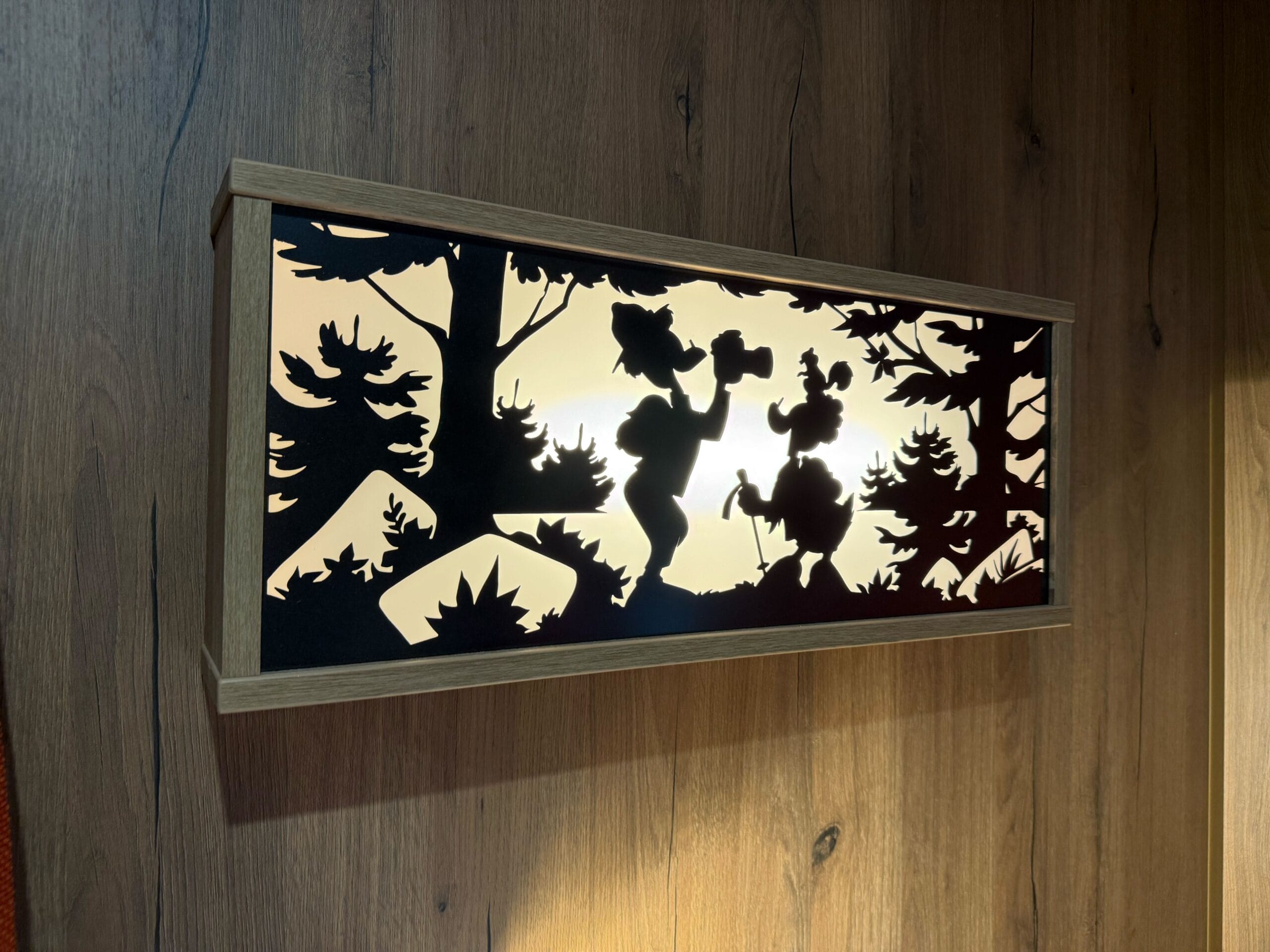 A rectangular light fixture with a cutout silhouetted scene of Donald (with a squirrel on his head) and another duck on a hike surrounded by trees.