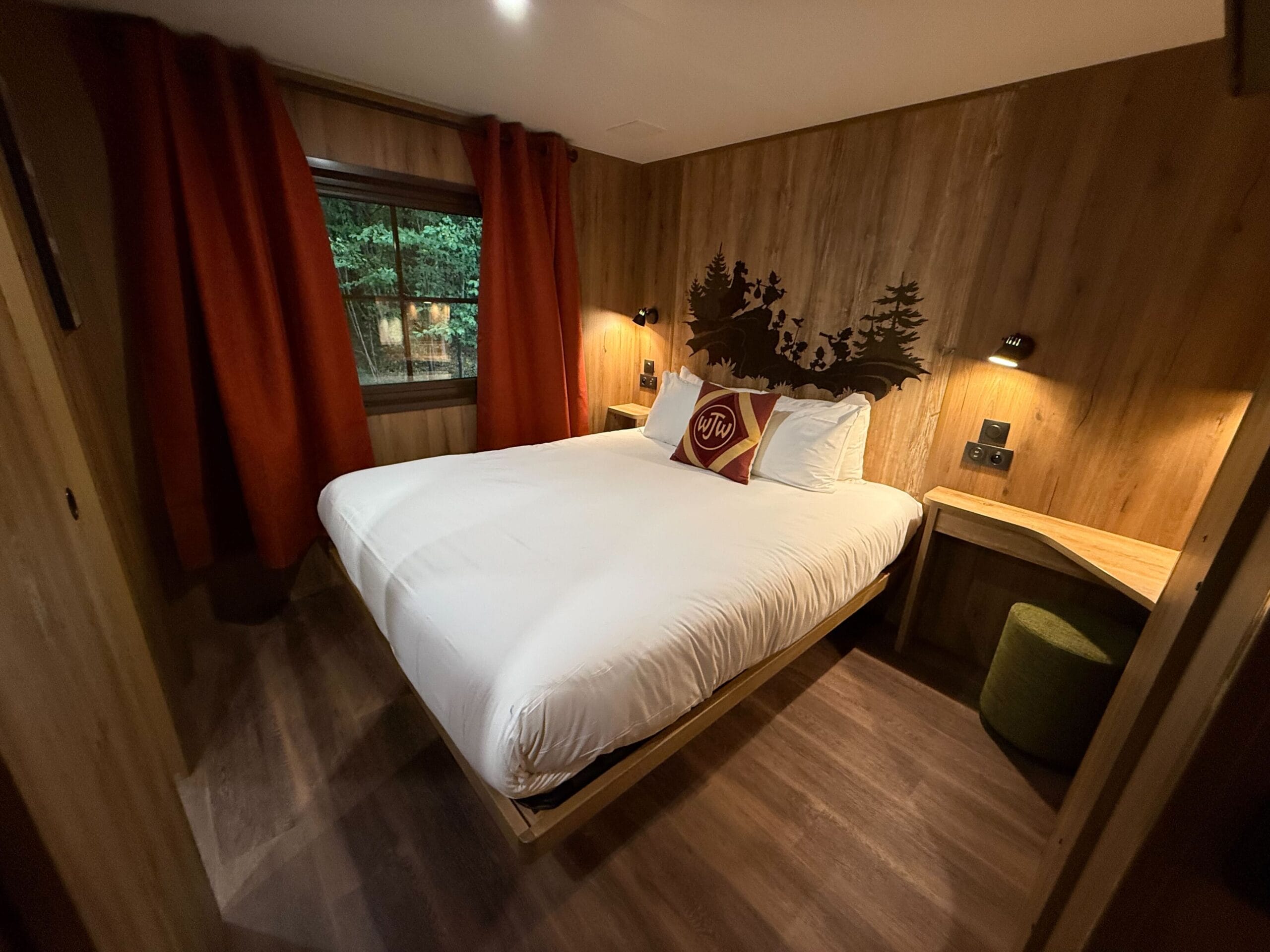 A cozy cabin bedroom at Disneyland Paris Davy Crockett Ranch with white linens, wood paneling, and reading lights, plus a Junior Woodchucks monogram pillow and woodland scene mural.
