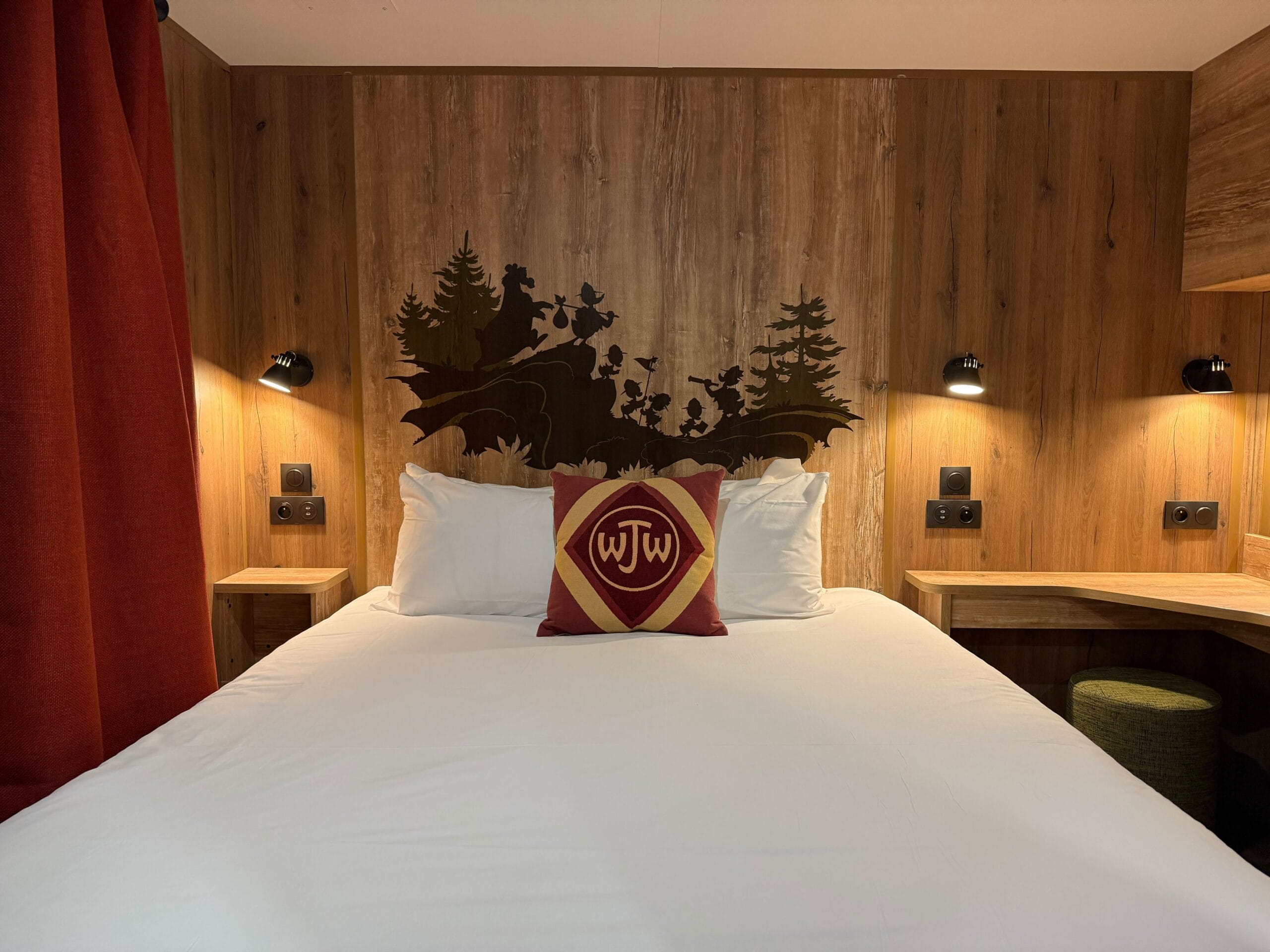 A cozy cabin bedroom at Disneyland Paris Davy Crockett Ranch with white linens, wood paneling, and reading lights, plus a Junior Woodchucks monogram pillow and woodland scene mural.