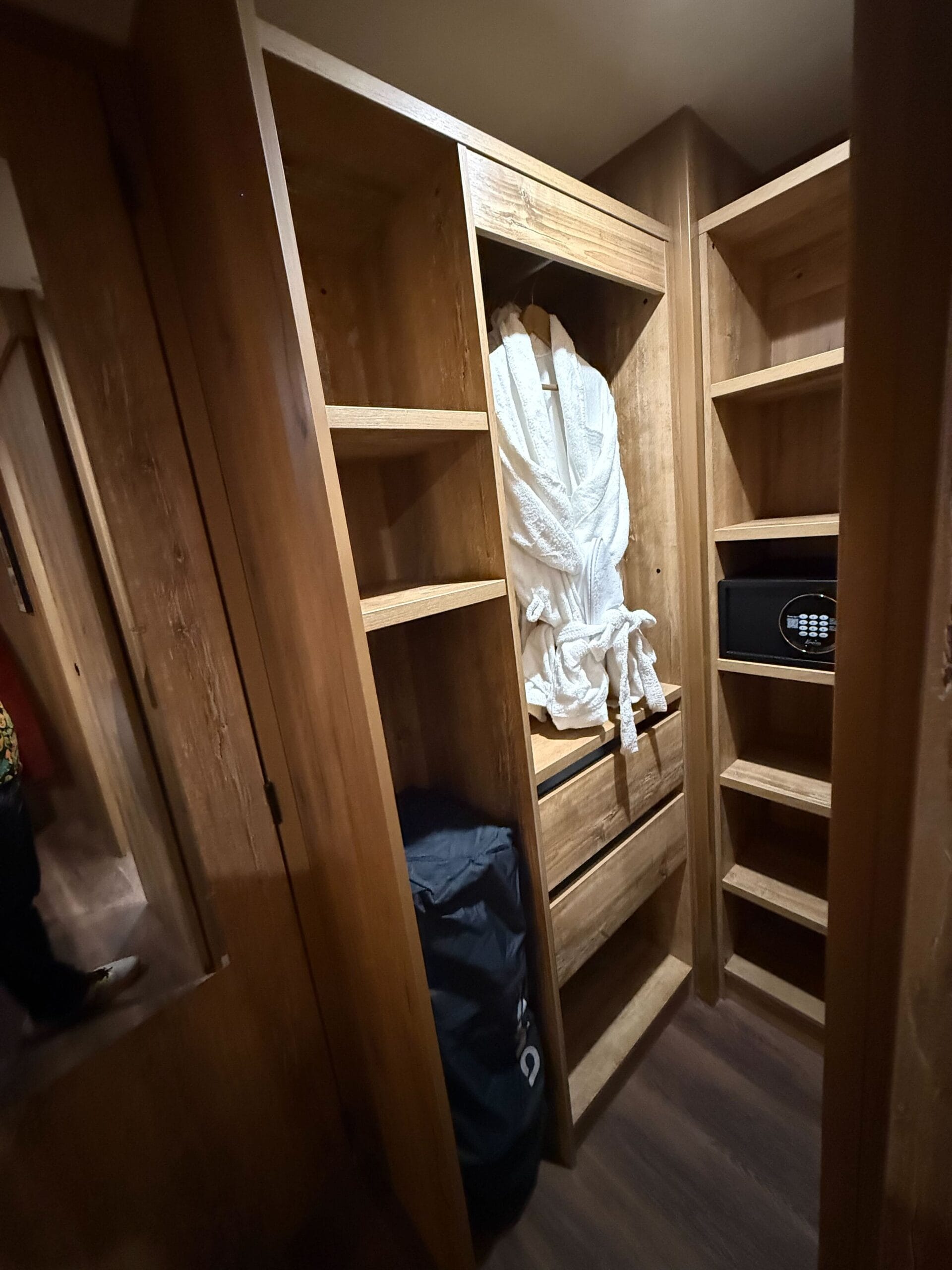 Wooden closet with shelves in a Davy Crockett Ranch cabin at Disneyland Paris, featuring robes, safe, and bag on dark wood floor.