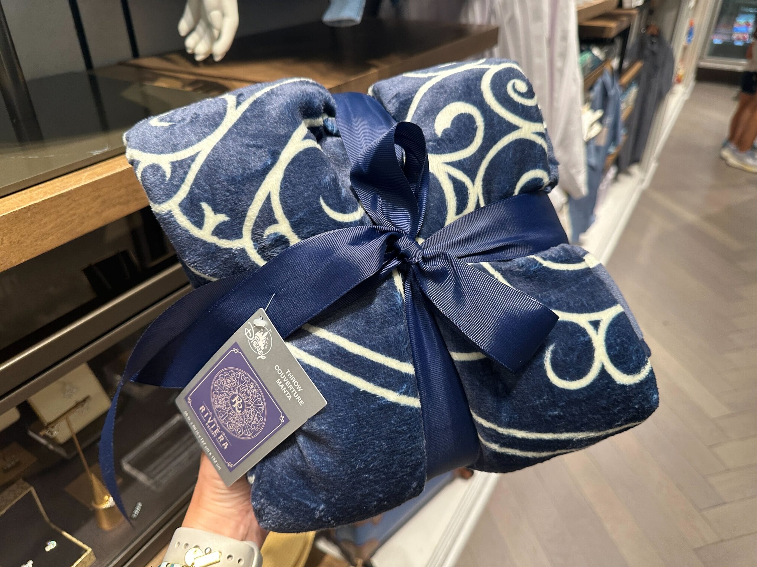 A hand displays a Disney's Riviera Resort blanket with elegant white swirls, ribbon, and gift tag—exclusive resort merchandise.