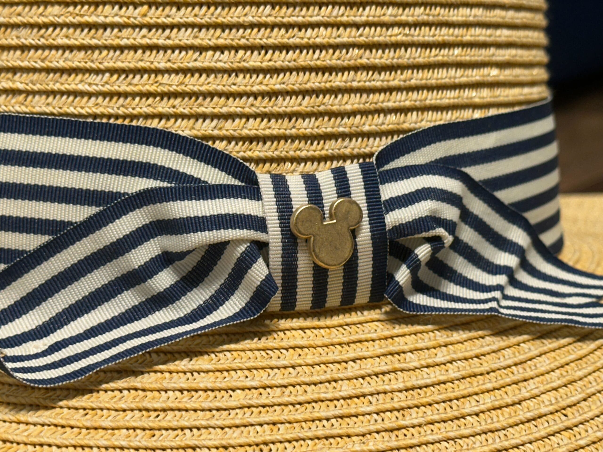 A straw hat featuring a navy striped bow, gold Mickey icon, and Riviera Resort style—evoking Disney resort elegance, not castle imagery.