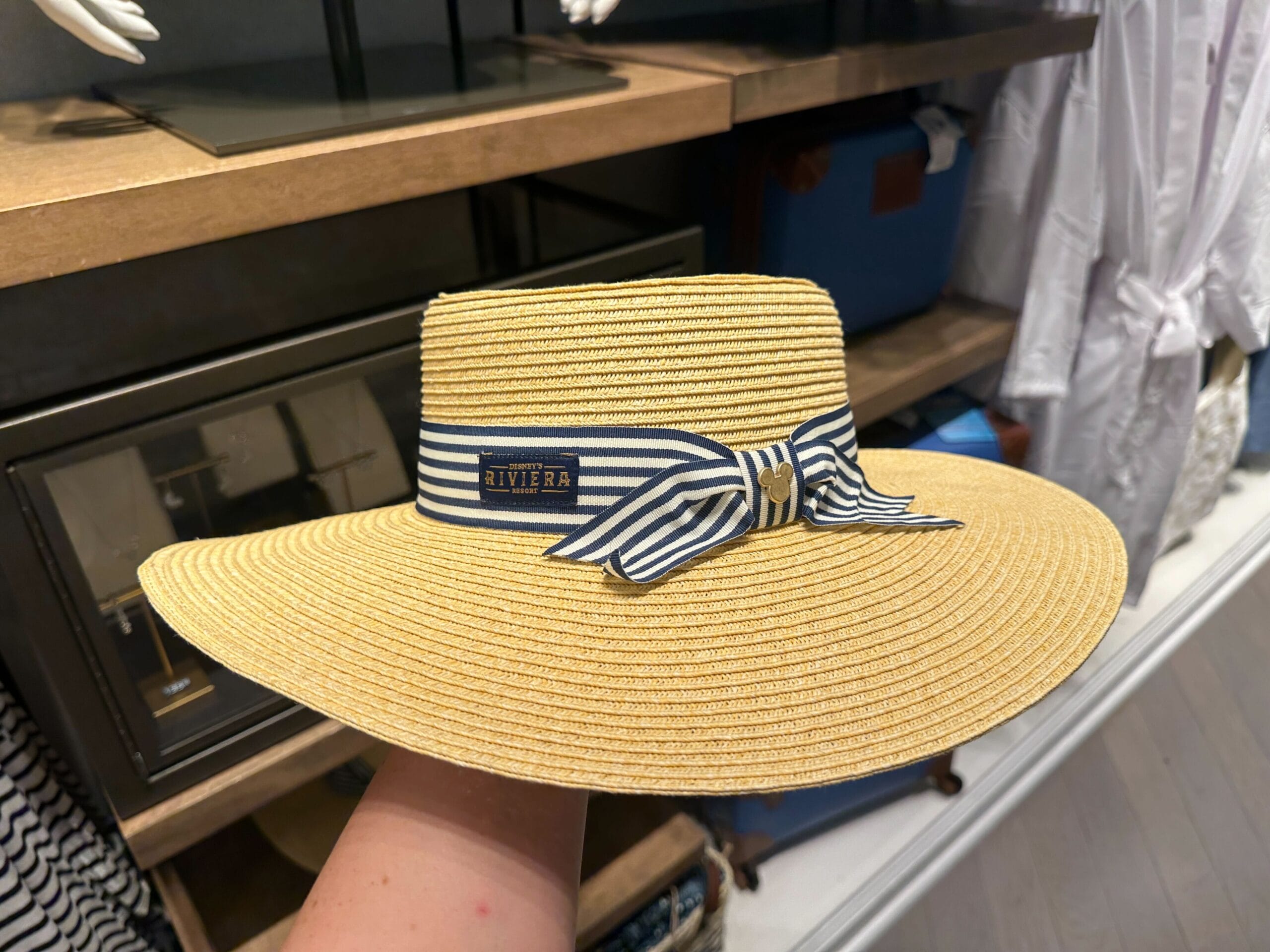 A hand displays a new Riviera Resort straw hat with a blue-and-white bow, exclusive to Disney Parks—no castle pictured here.