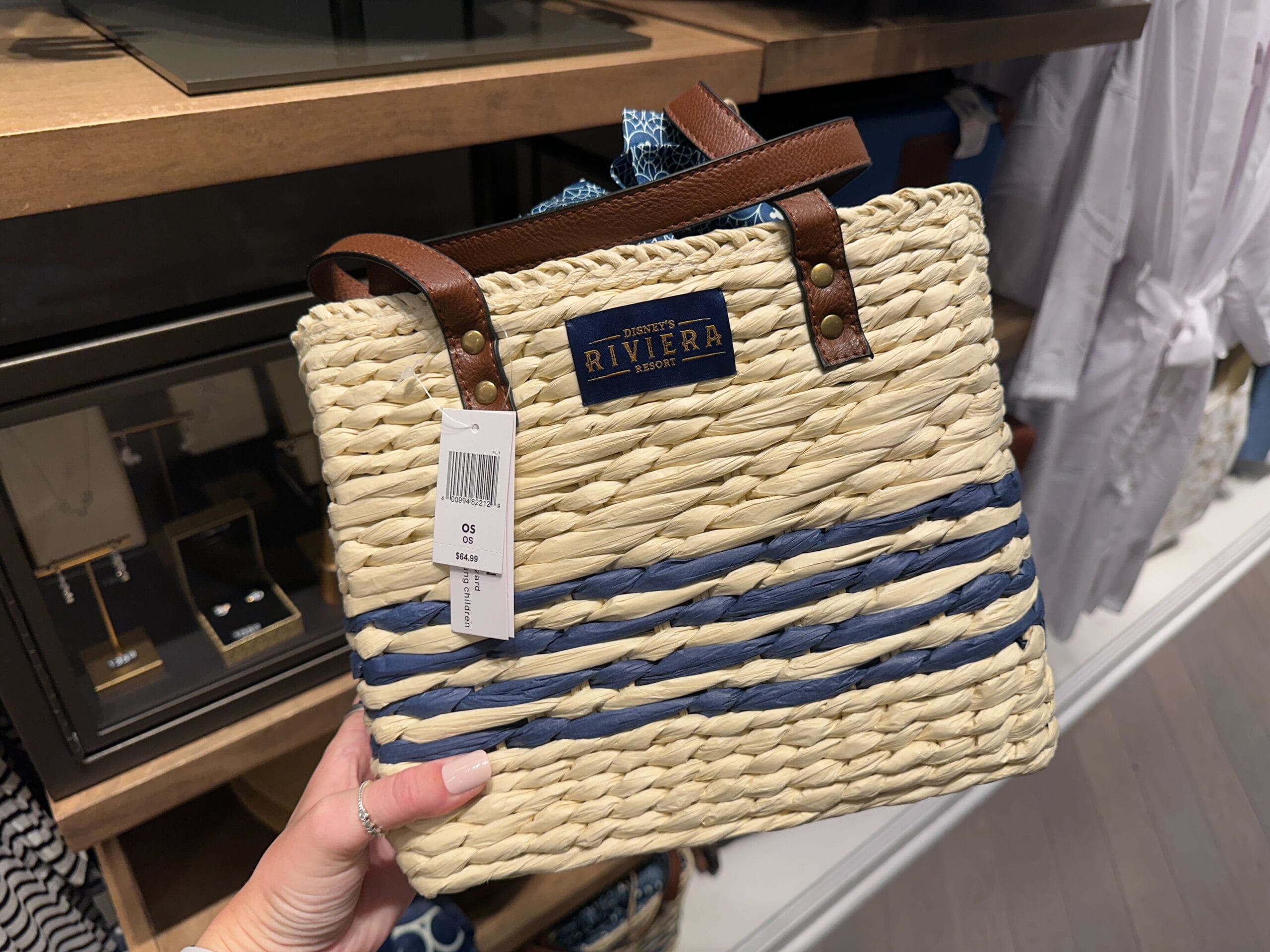 A hand displays a straw tote bag with blue stripes and brown handles, featuring a Disney's Riviera Resort label in a shop at Walt Disney World.