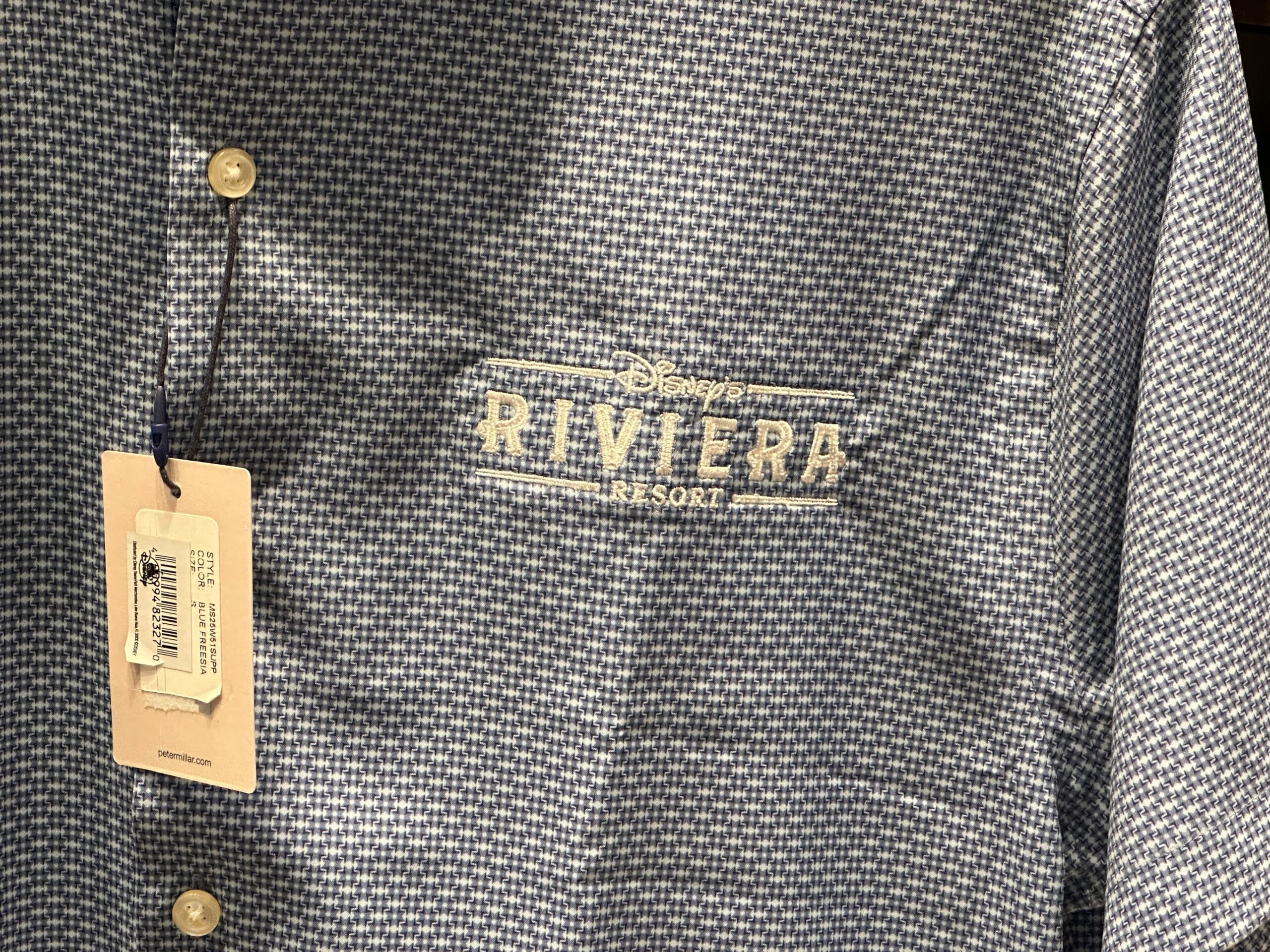 Close-up of a blue patterned shirt with "Disney Riviera Resort" embroidered, new in the parks, beige tag on buttonhole. No castle shown.
