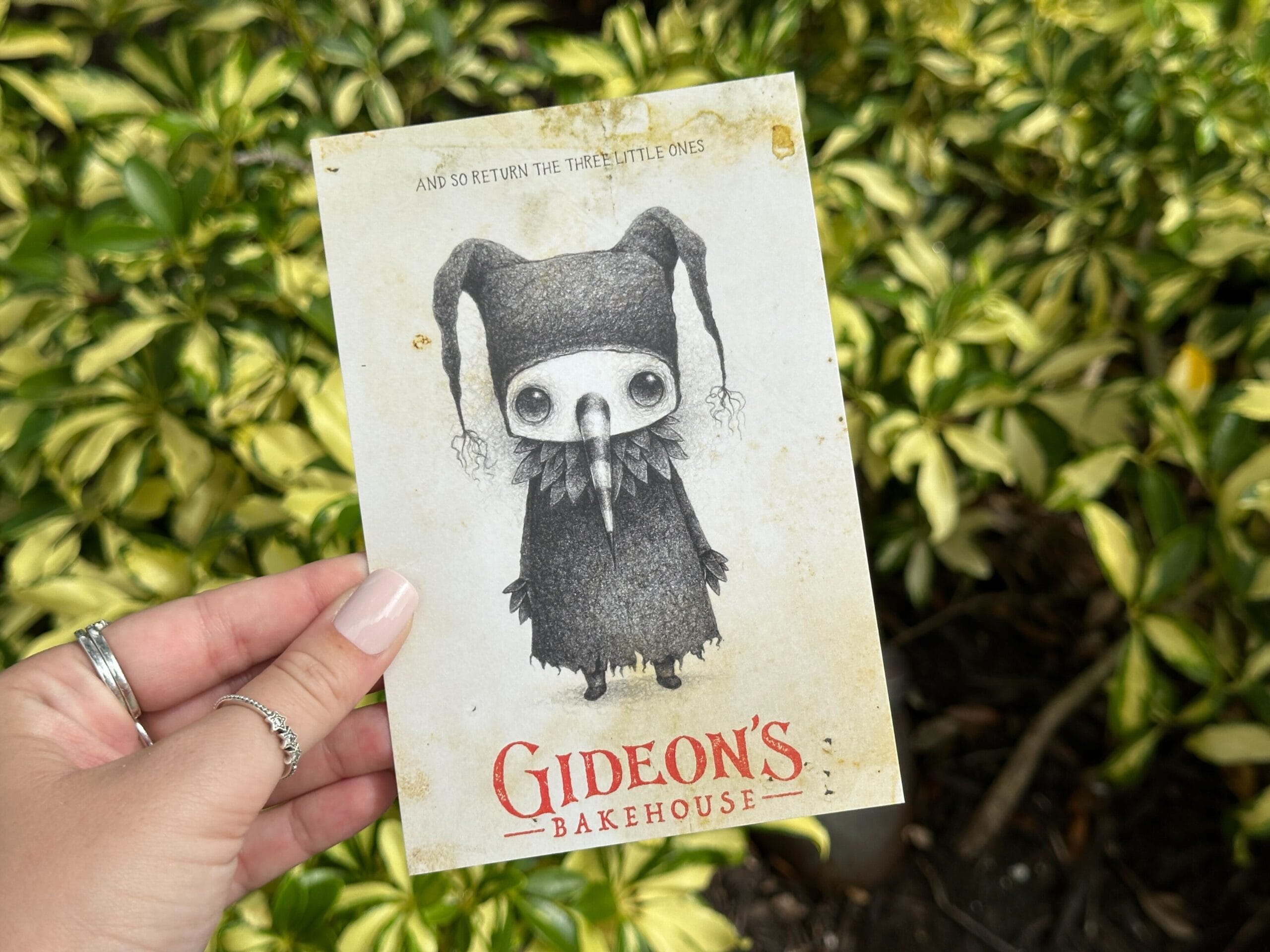A hand holds a Gideon's Bakehouse card featuring a sketch of a spooky jester character against a leafy background.