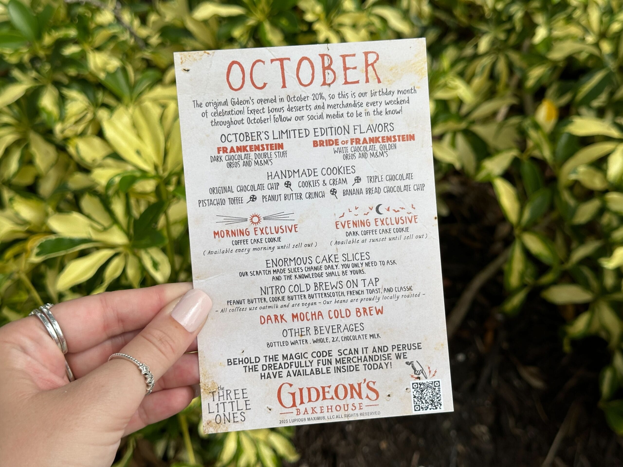 A hand holds a Gideon's Bakehouse October menu in front of green and yellow leafy plants.