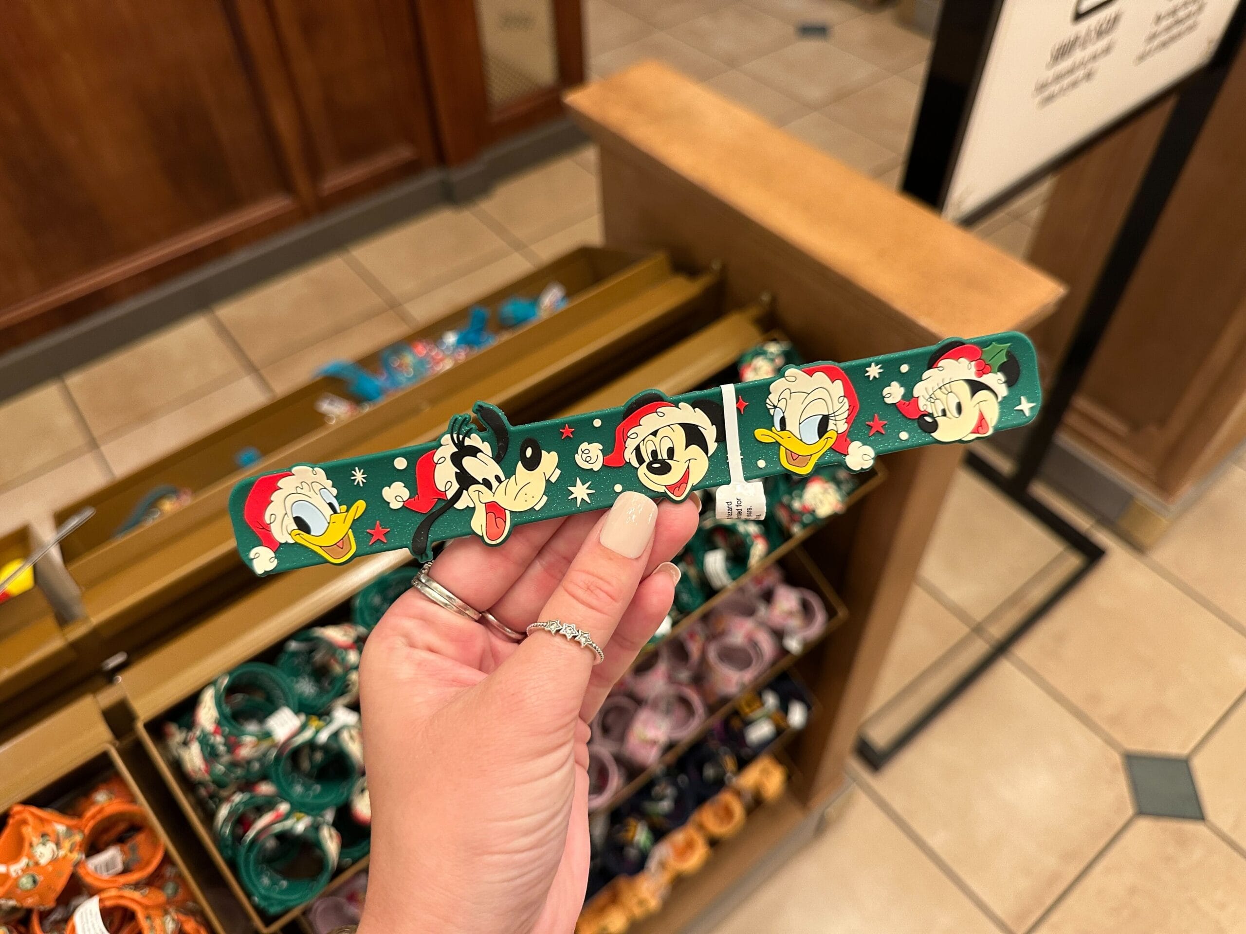 A hand displays a green Walt Disney World Christmas wristband featuring Donald, Goofy, Mickey, and Pluto in festive hats.