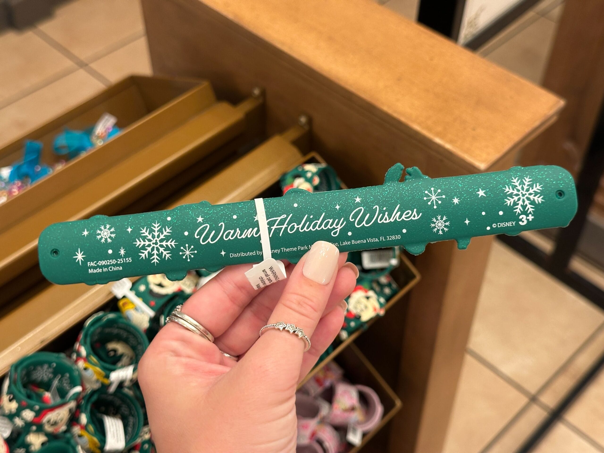 A hand displays a green Walt Disney World holiday slap bracelet reading "Warm Holiday Wishes" with snowflakes, exclusive to WDW.