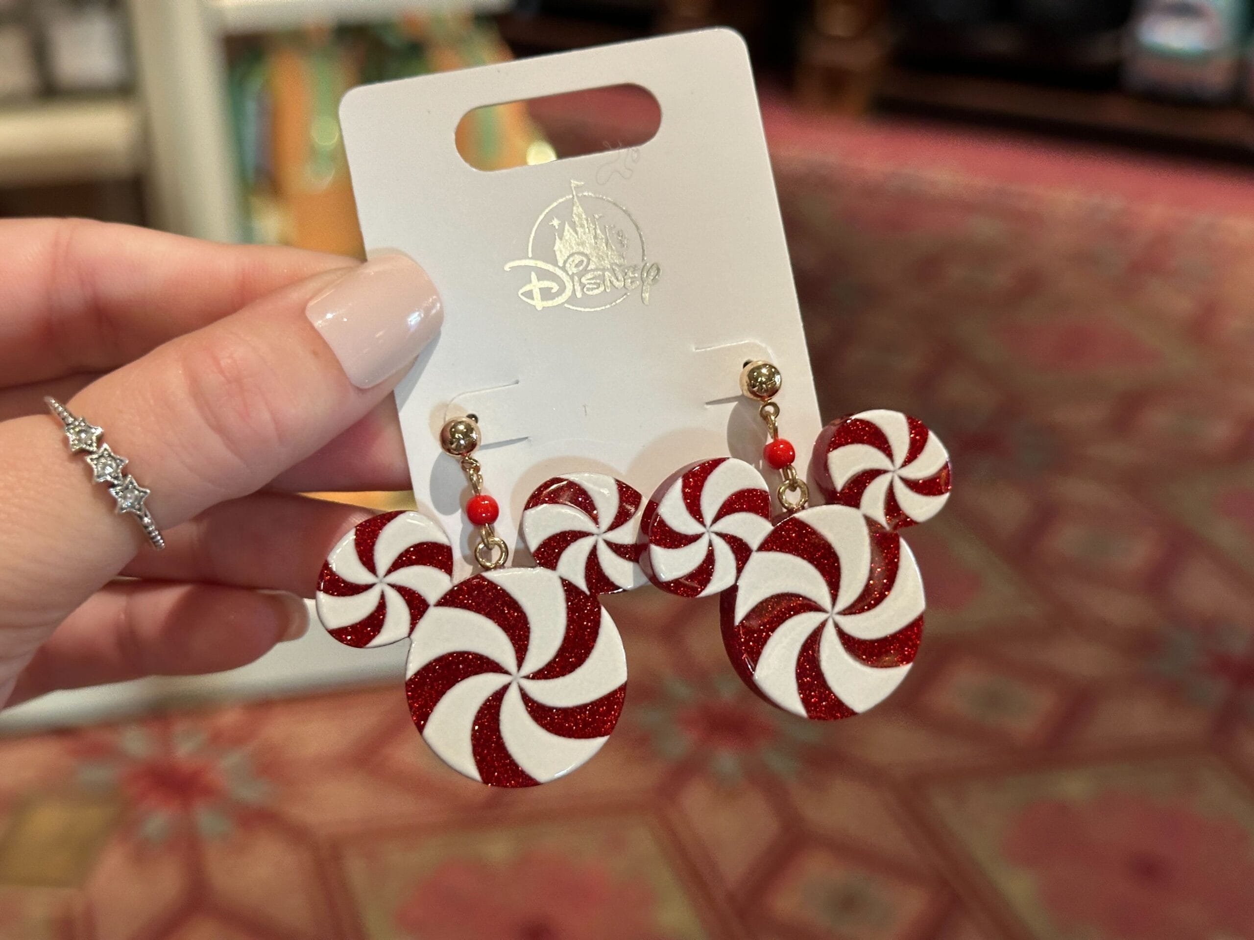 Hand holding red and white glitter peppermint swirl earrings with gold accents, festive 2025 Walt Disney World Christmas merchandise.