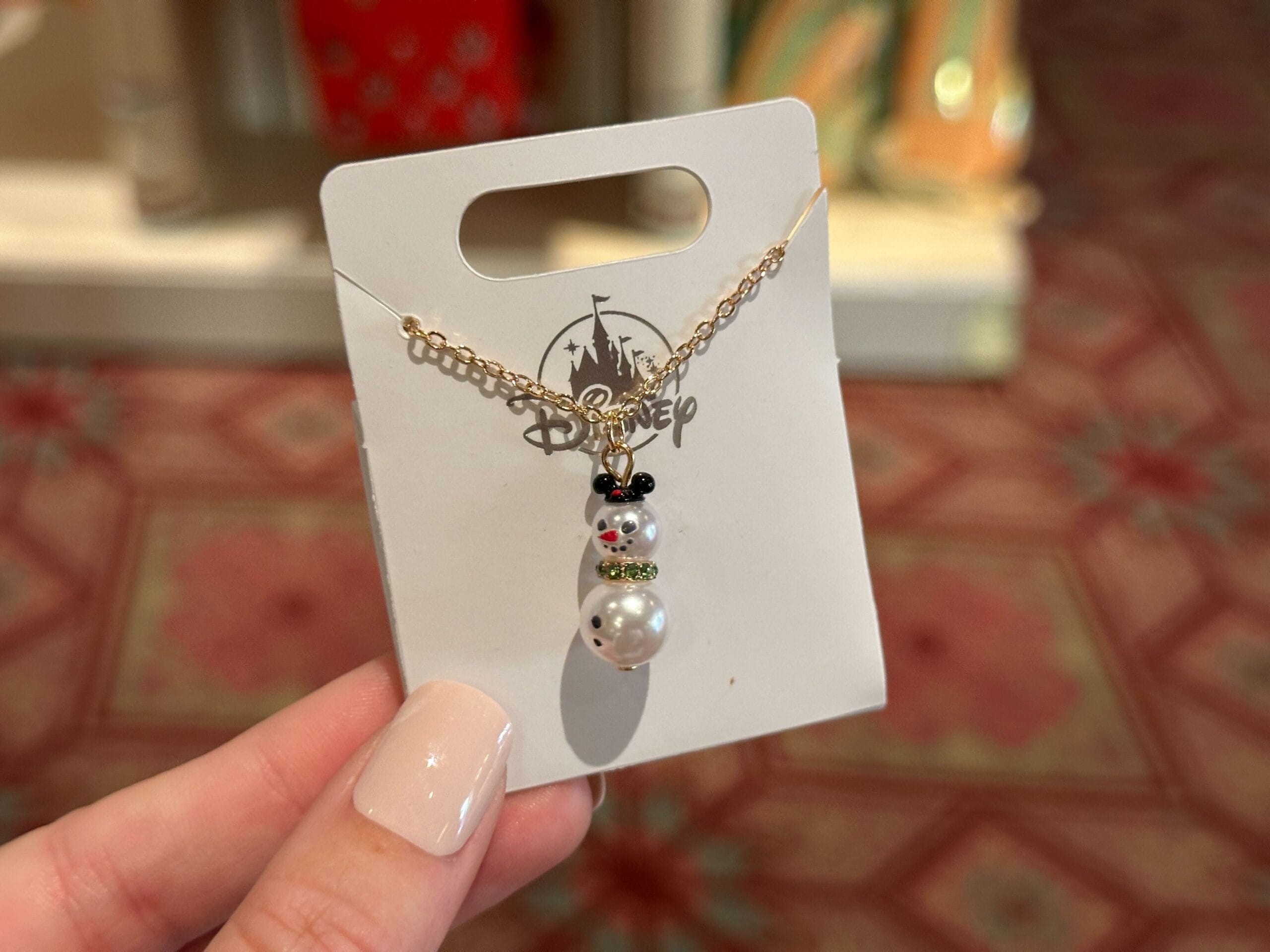 Hand holding a 2025 snowman Mickey Mouse necklace, festive holiday merchandise on Walt Disney World backing near castle motif.
