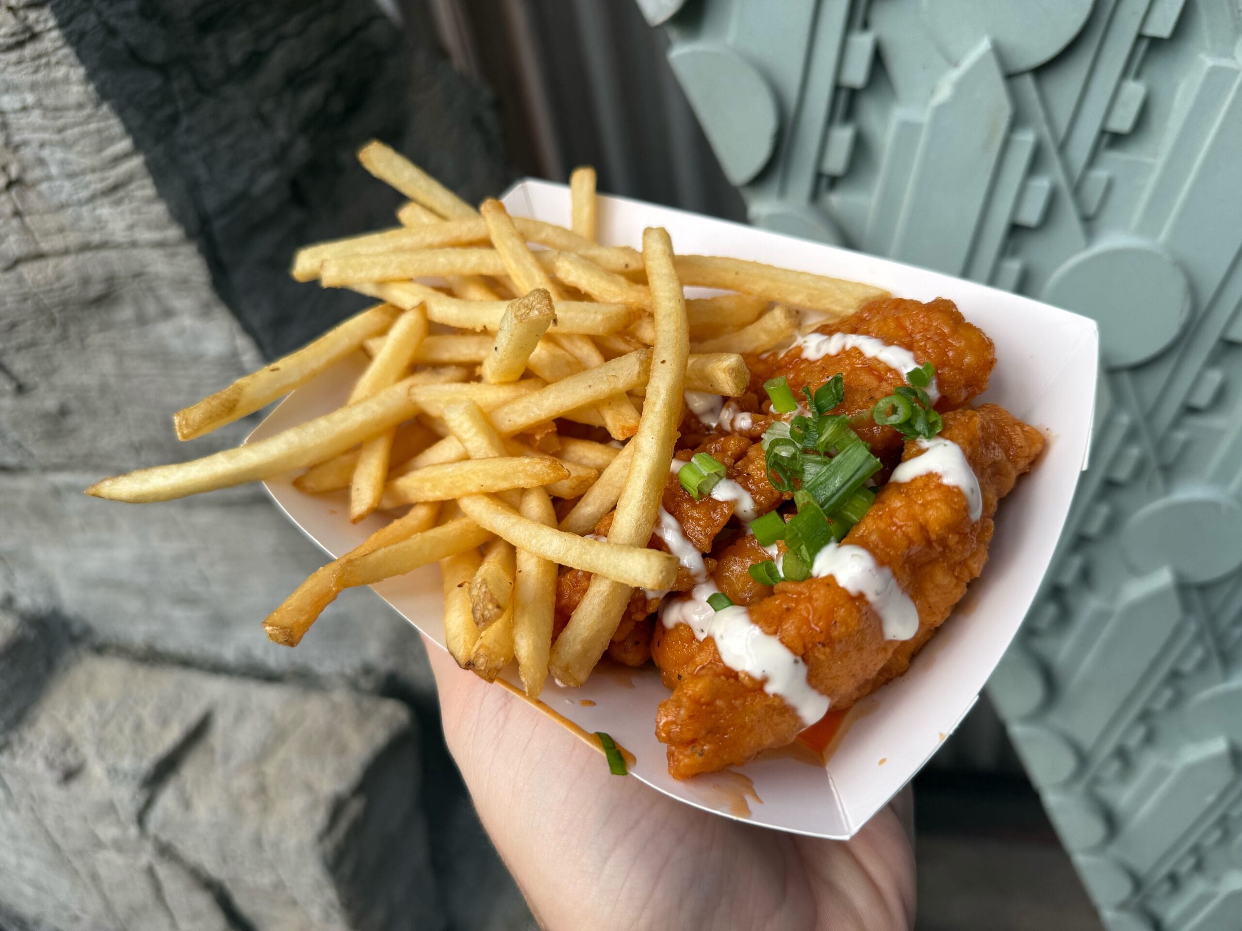 A hand holds saucy Honey Buffalo Chicken Strips and crispy fries at Disney’s Hollywood Studios, garnished with green onions.