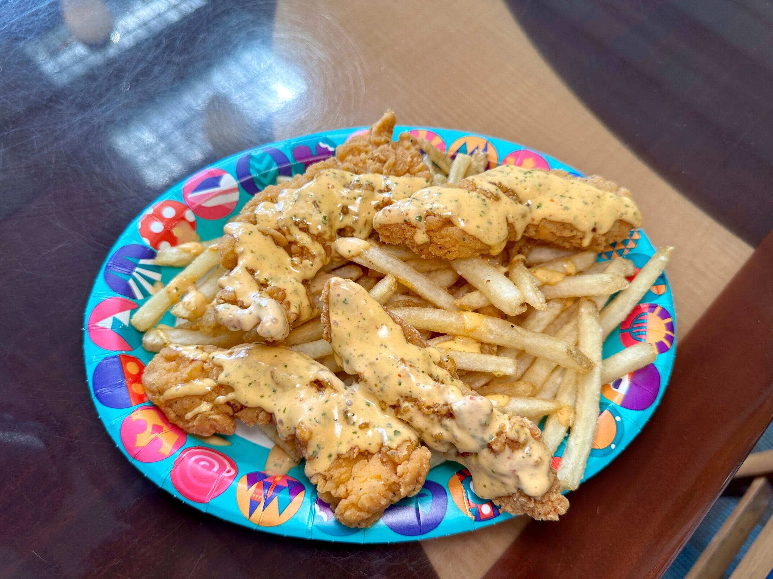 Spicy Ranch Chicken Strips with creamy sauce over French fries on a colorful paper plate, like a snack from Disney parks.