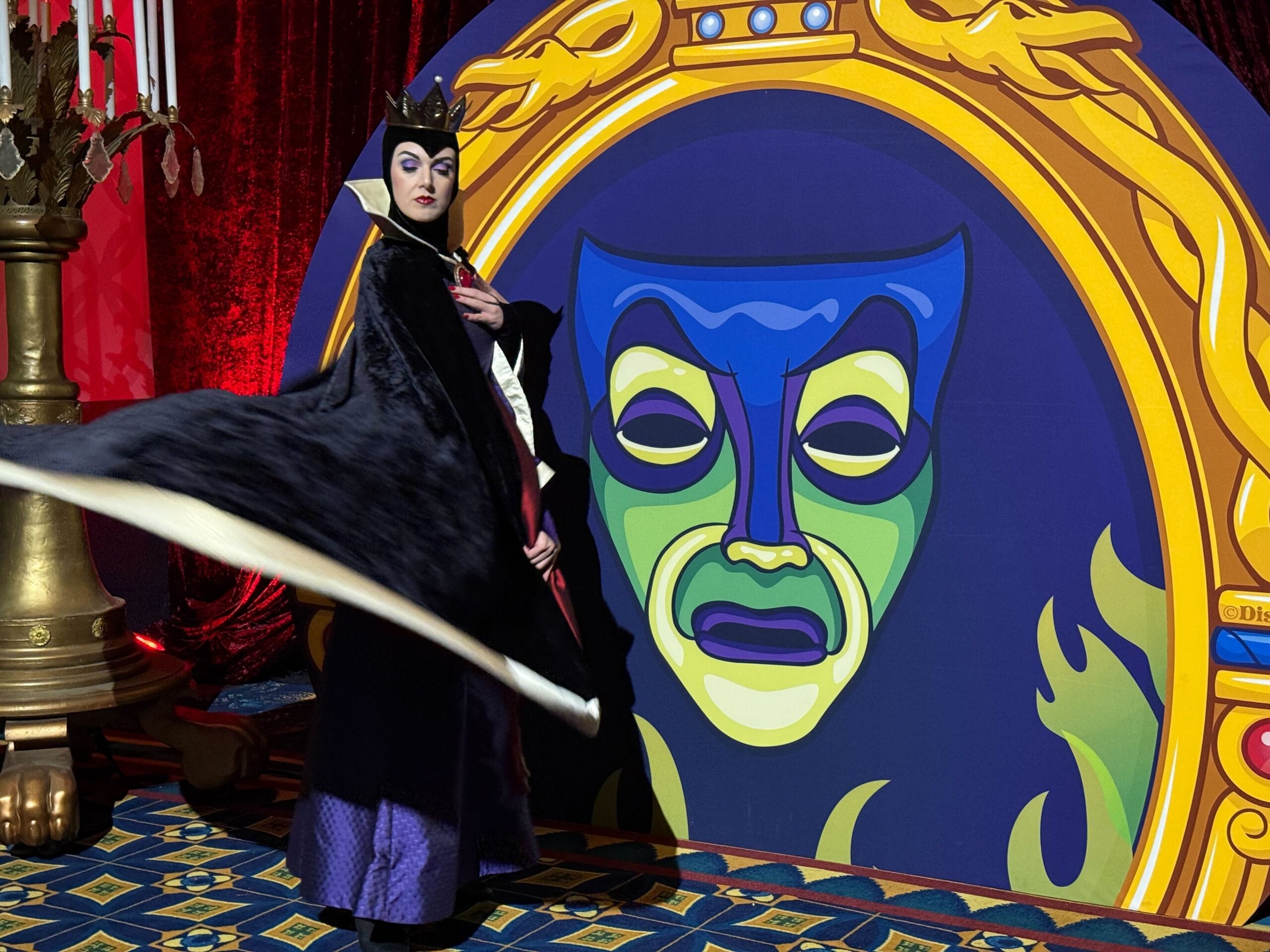 A person dressed as the Evil Queen poses dramatically beside a large, colorful Magic Mirror at Trader Sam's.