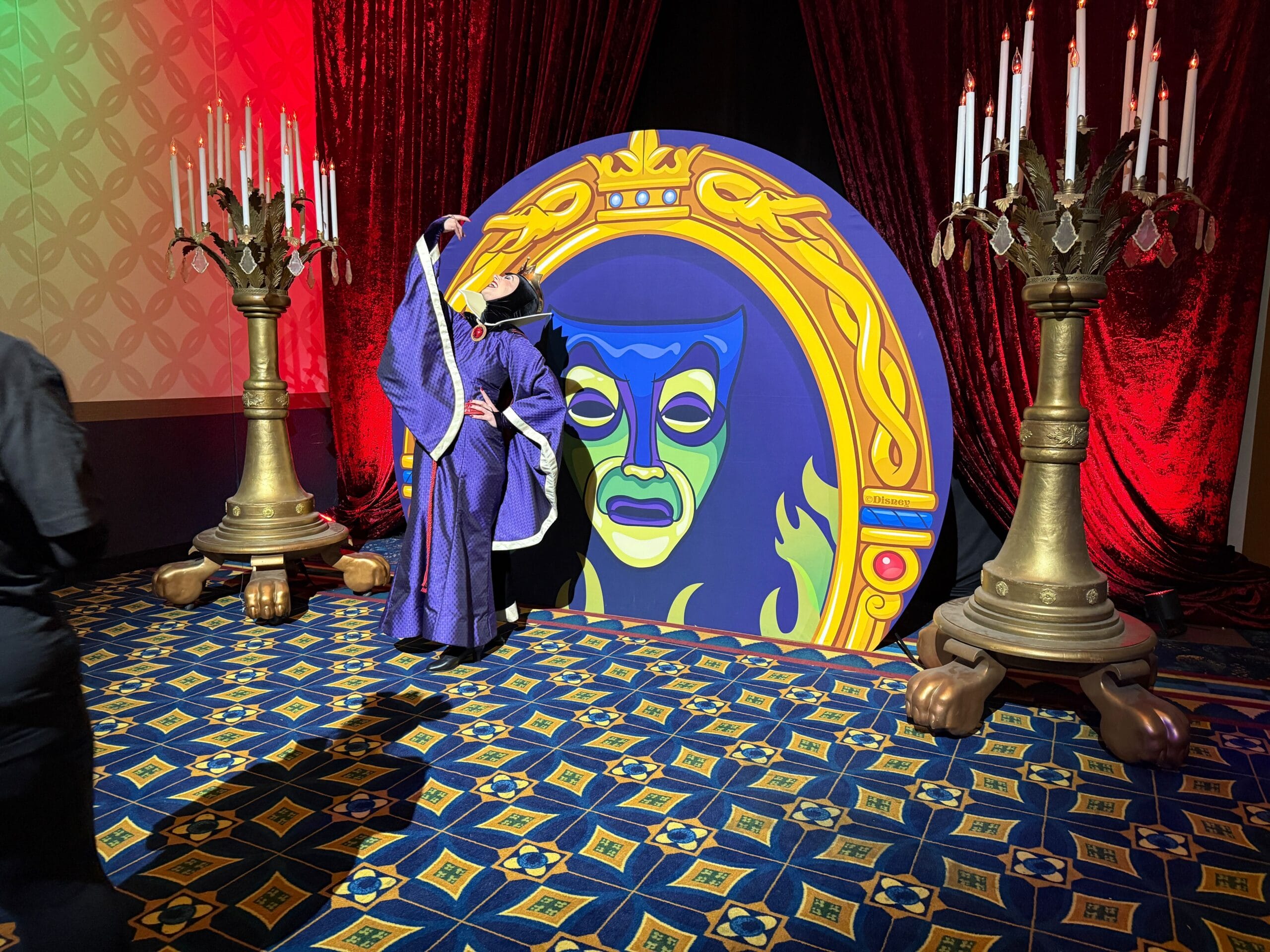 A person in a purple villain costume poses by Trader Sam's magic mirror with a green face, beside ornate candle stands.