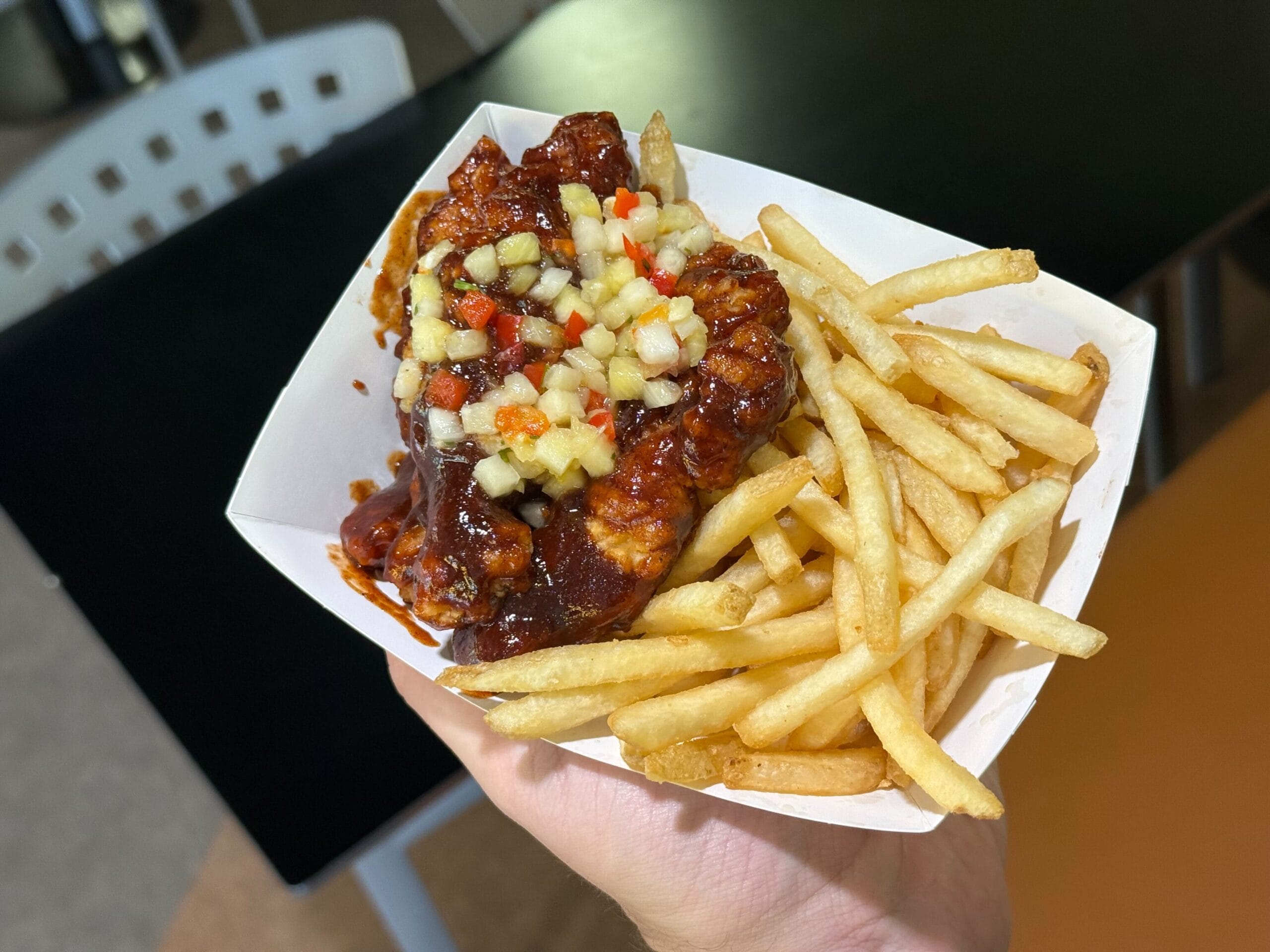 A hand holds a tray of French fries and saucy Jerk BBQ Chicken topped with diced veggies at a Disney park food stand.