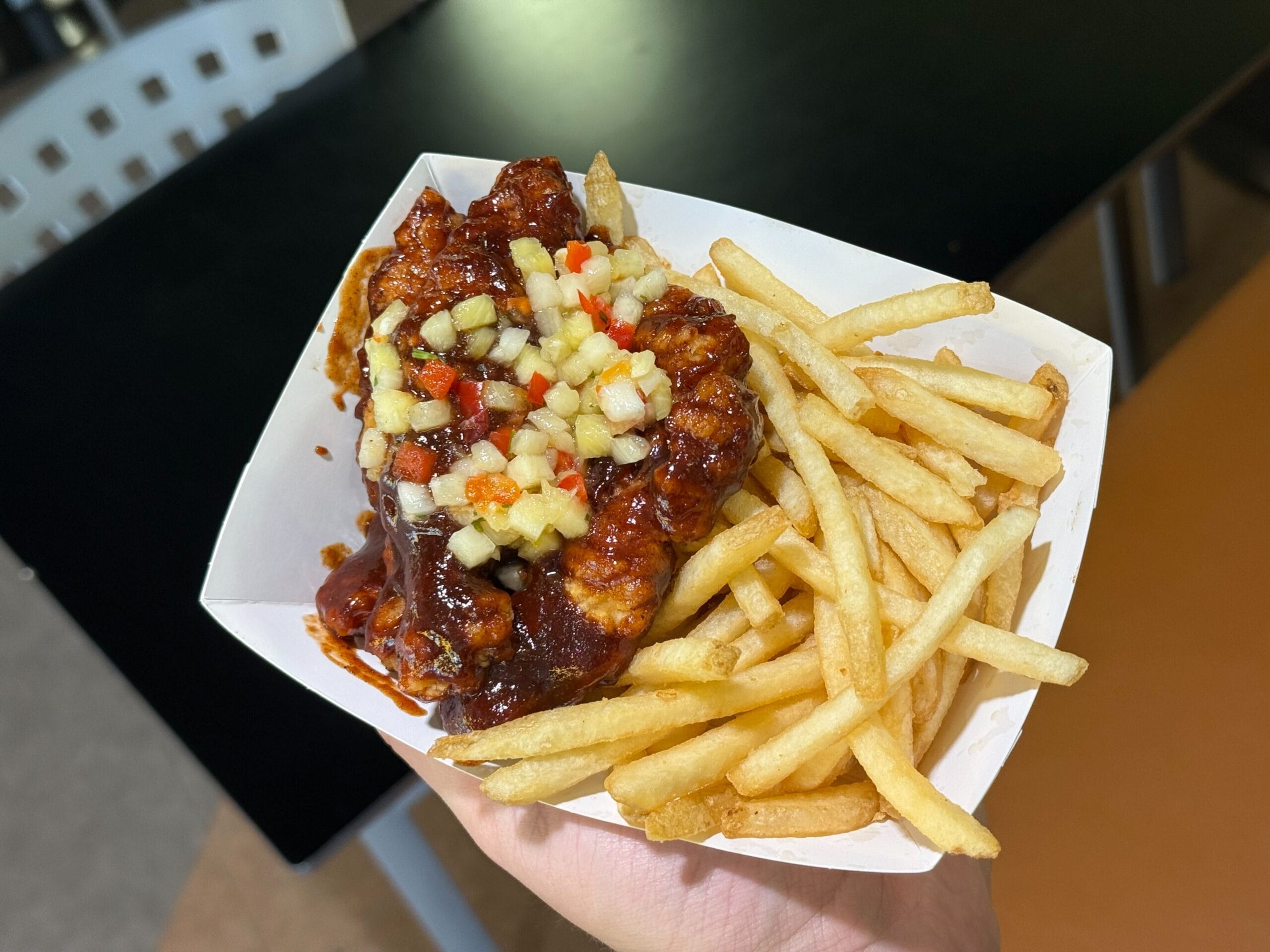 A hand holds a Disney Parks food tray filled with Jerk BBQ chicken strips topped with pineapple salsa and served with fries.