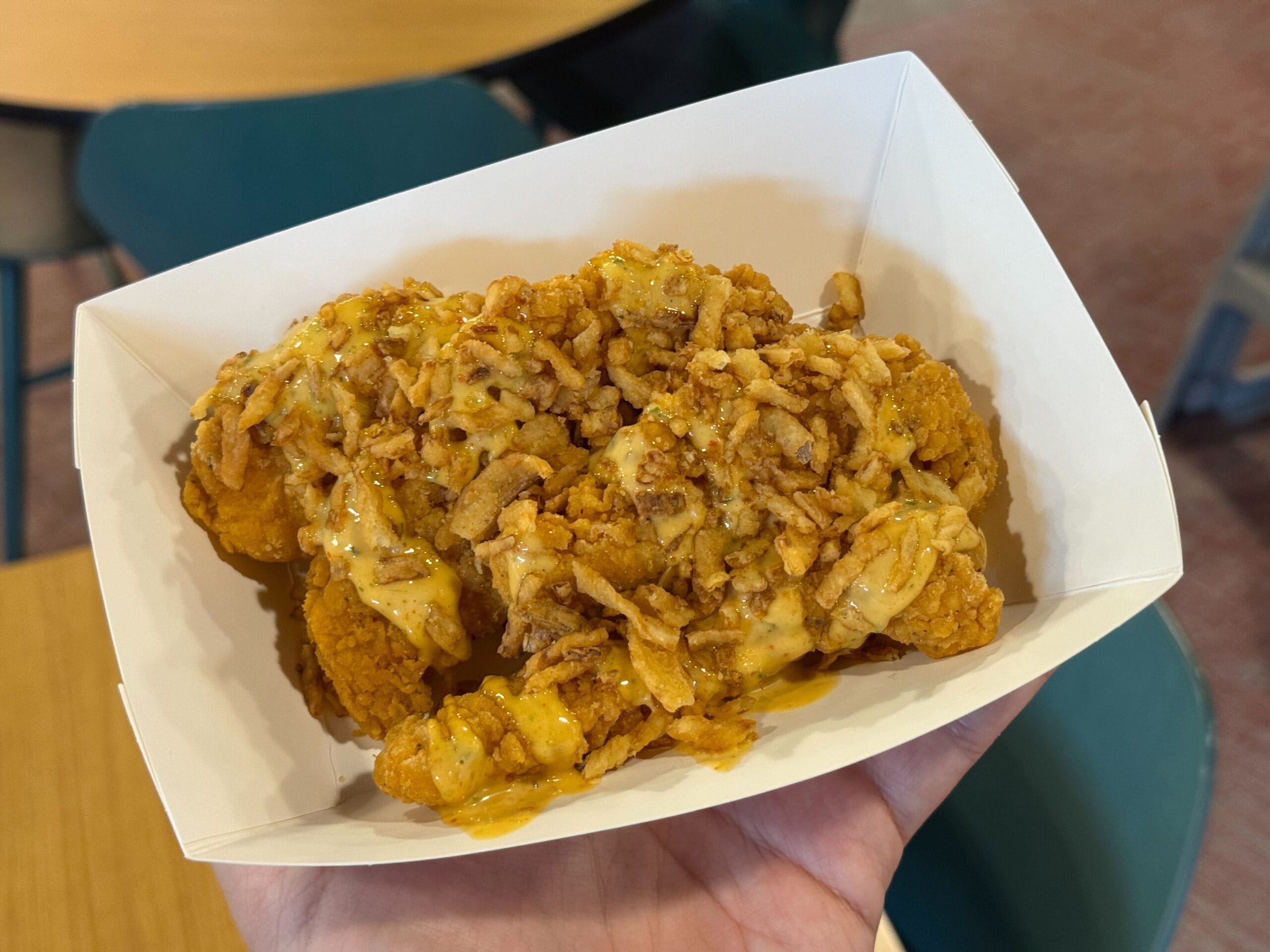 A hand holds a tray of Chicken Strips with sweet chili aïoli and crispy onions, a popular Disney Parks snack for on-the-go guests.