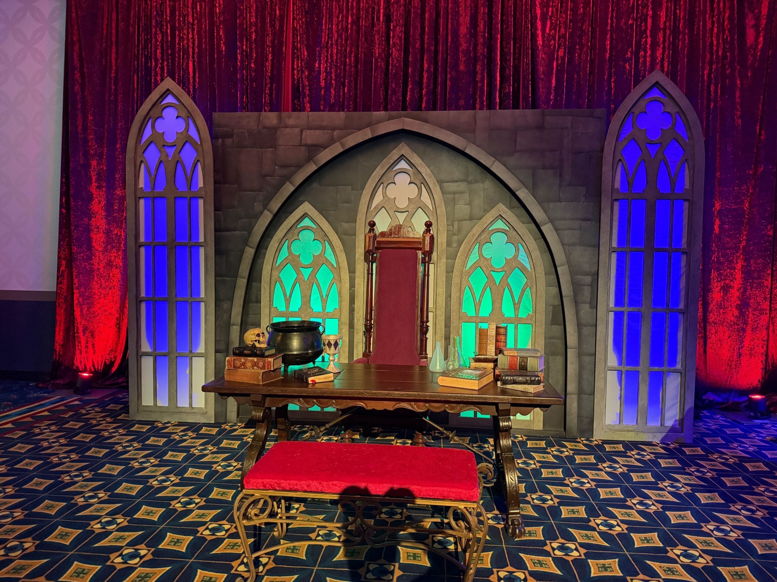 A throne sits behind a table with books, props, and an Evil Queen Mug, framed by Gothic arches and colorful lighting.