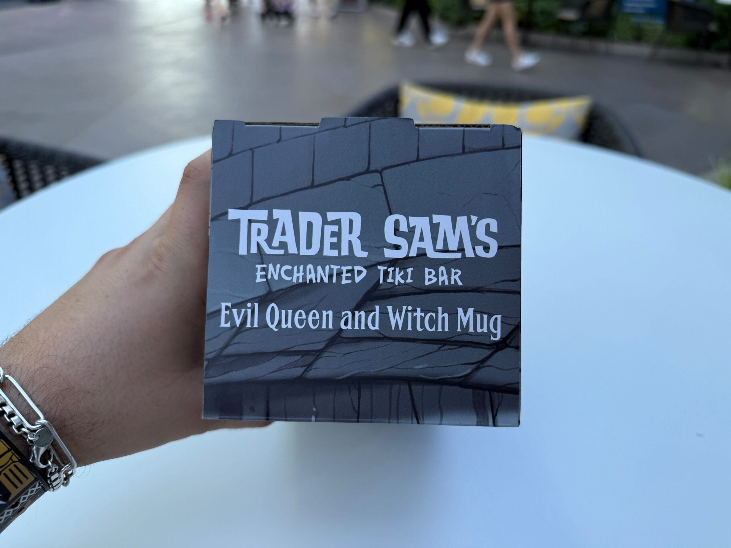 Hand holding a Trader Sam's Evil Queen Mug box on an outdoor table at the Disneyland Hotel.