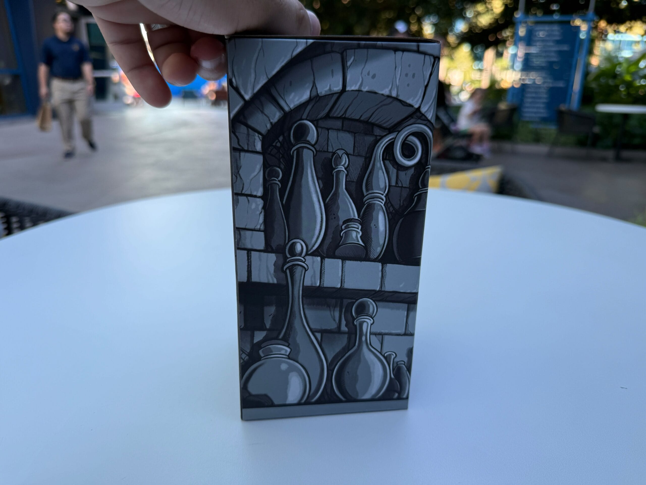 A hand holds a box with black and white art of potion bottles, inspired by Trader Sam's, on a round white table outdoors.