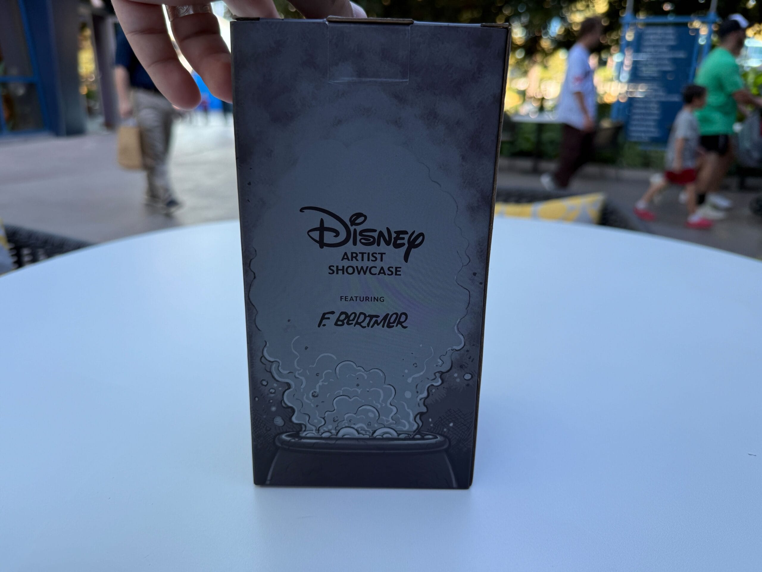A hand holds a Disney Artist Showcase box and an Evil Queen Mug on a white table at the Disneyland Hotel.