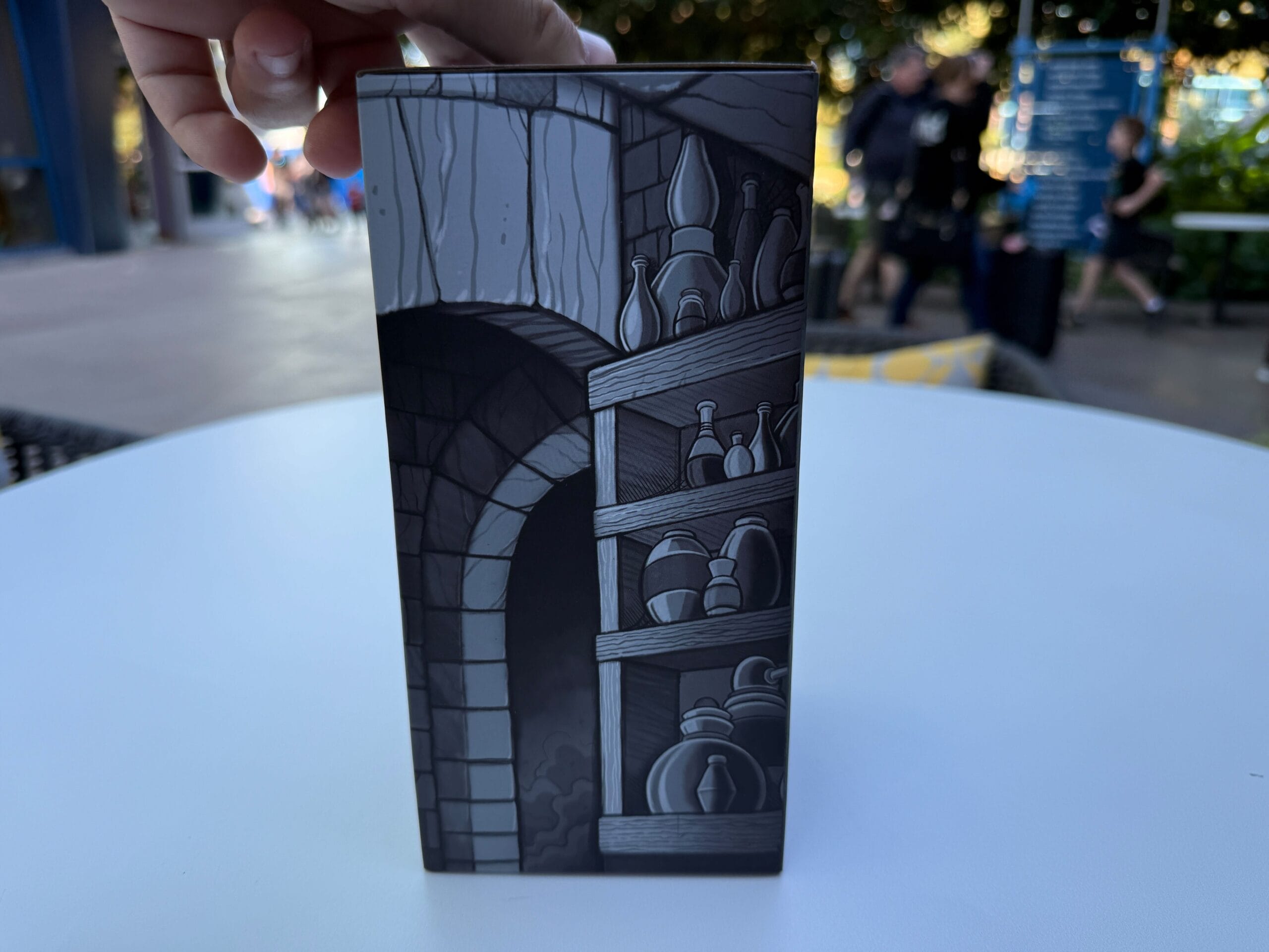 Hand holding a box with illustrated shelves of bottles at Trader Sam's; people blurred in background.