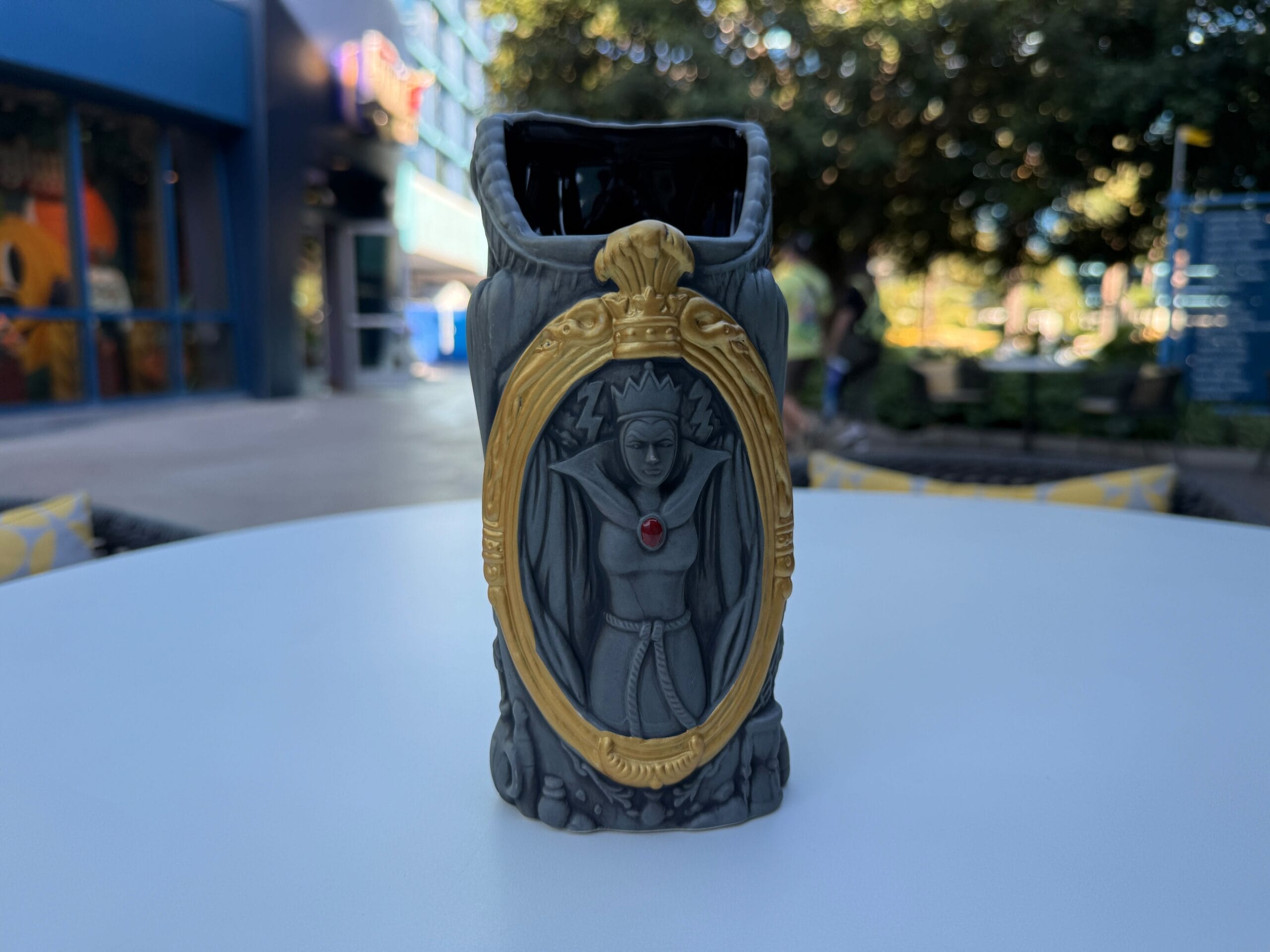 A decorative Evil Queen Mug with a gold-framed image and red gem sits on a white table outdoors.